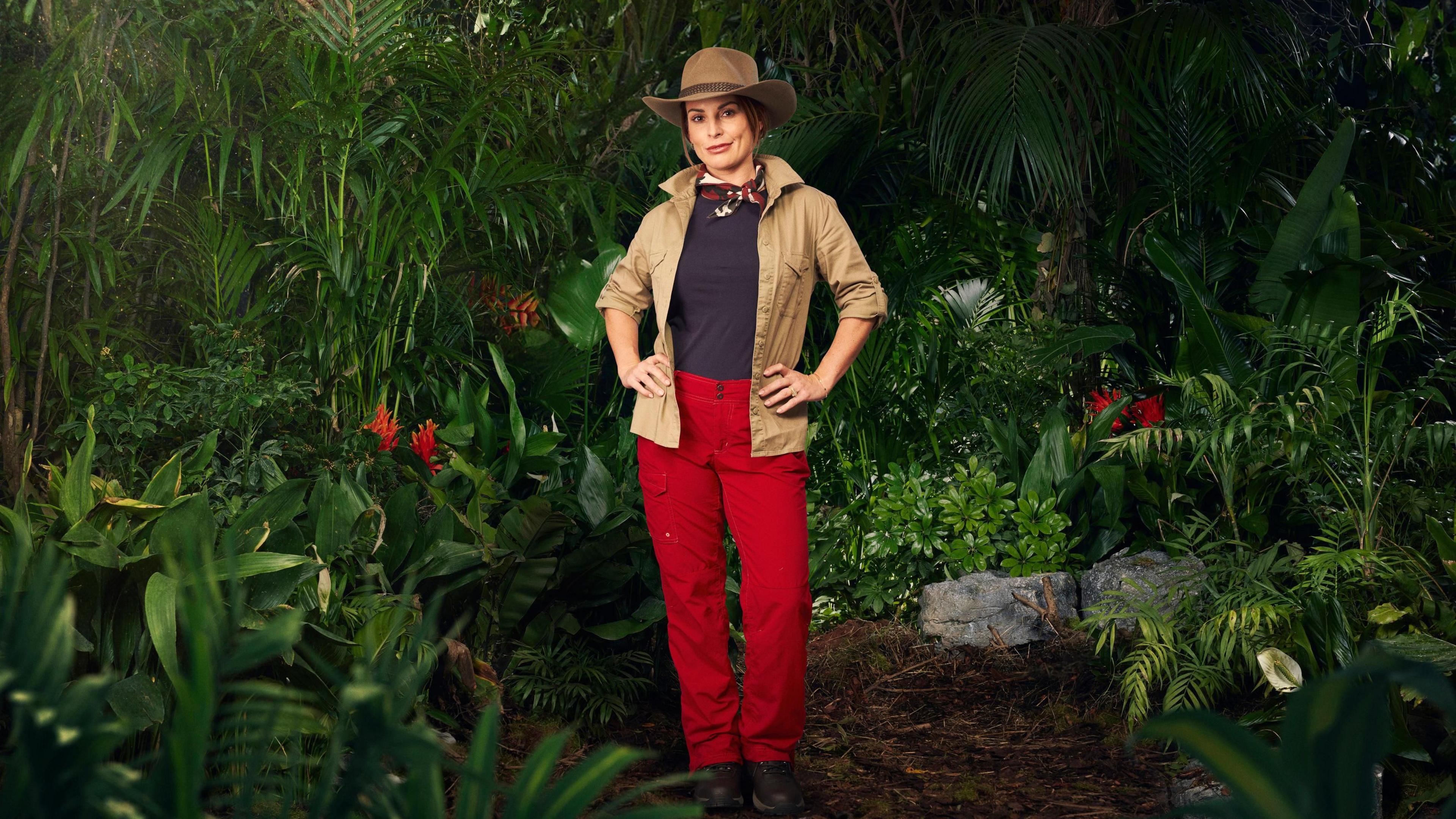 Coleen Ronney wears a beigh flak jacket, red trousers and  a necktie and is pouting directly at the camera with her hands on her hips. In the background are jungle plants and red flowers. 