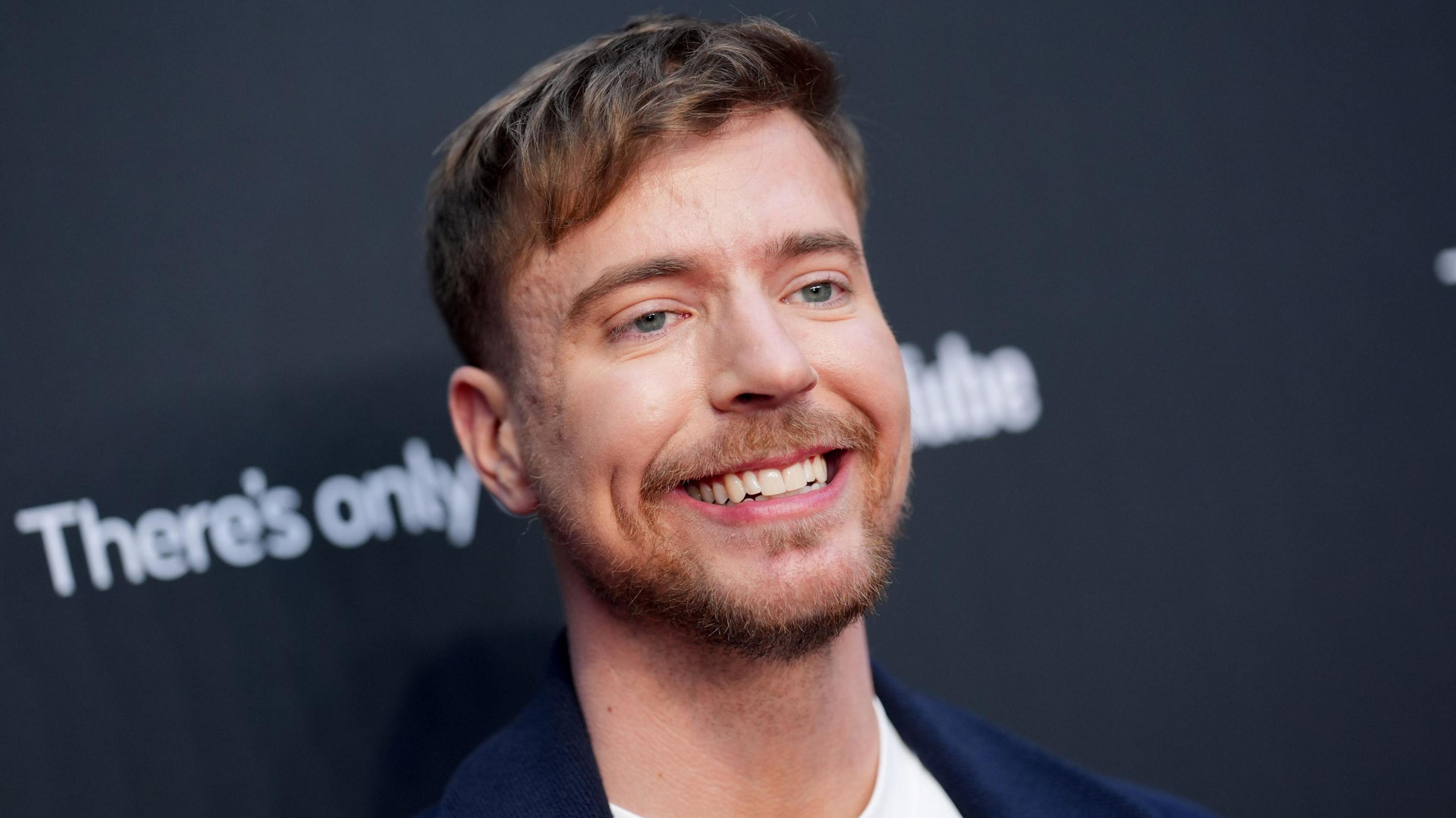 MrBeast pictured in New York in May 2025. The creator has blue eyes, short brown hair and a short beard. He smiles, wearing a dark blue blazer over a white T-shirt while posing for photographers in front of black hoarding.
