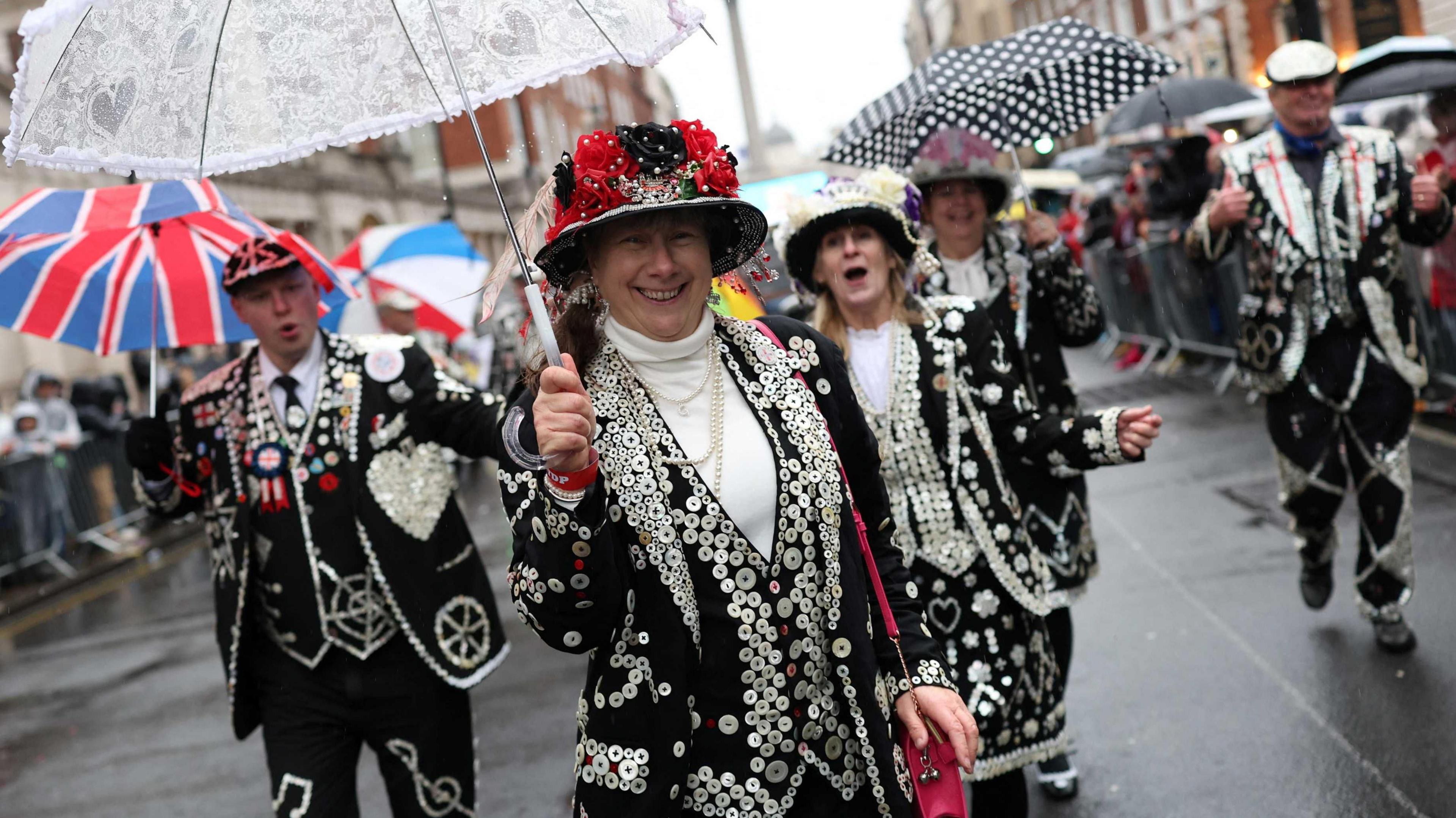 Four Pearly Kings and Queens in their distinctive, decorated clothing with umbrellas in hand take part in the New Year's Day parade. They are in the street and crowds of people can be seen in the background, standing behind barriers on the pavement