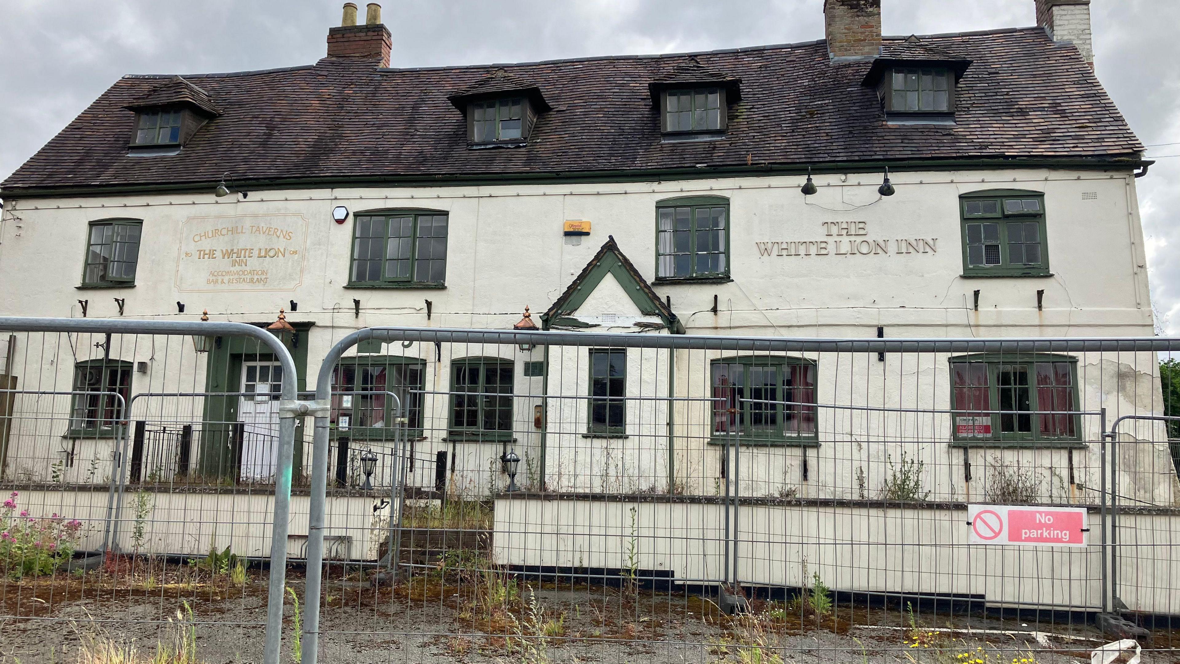 The exterior of a pub, there is a metal fence in the foreground with a no parking sign. The pub is white with green window frames, and a sign that says "The White Lion Inn". There are weeds growing out of the ground in front of the building