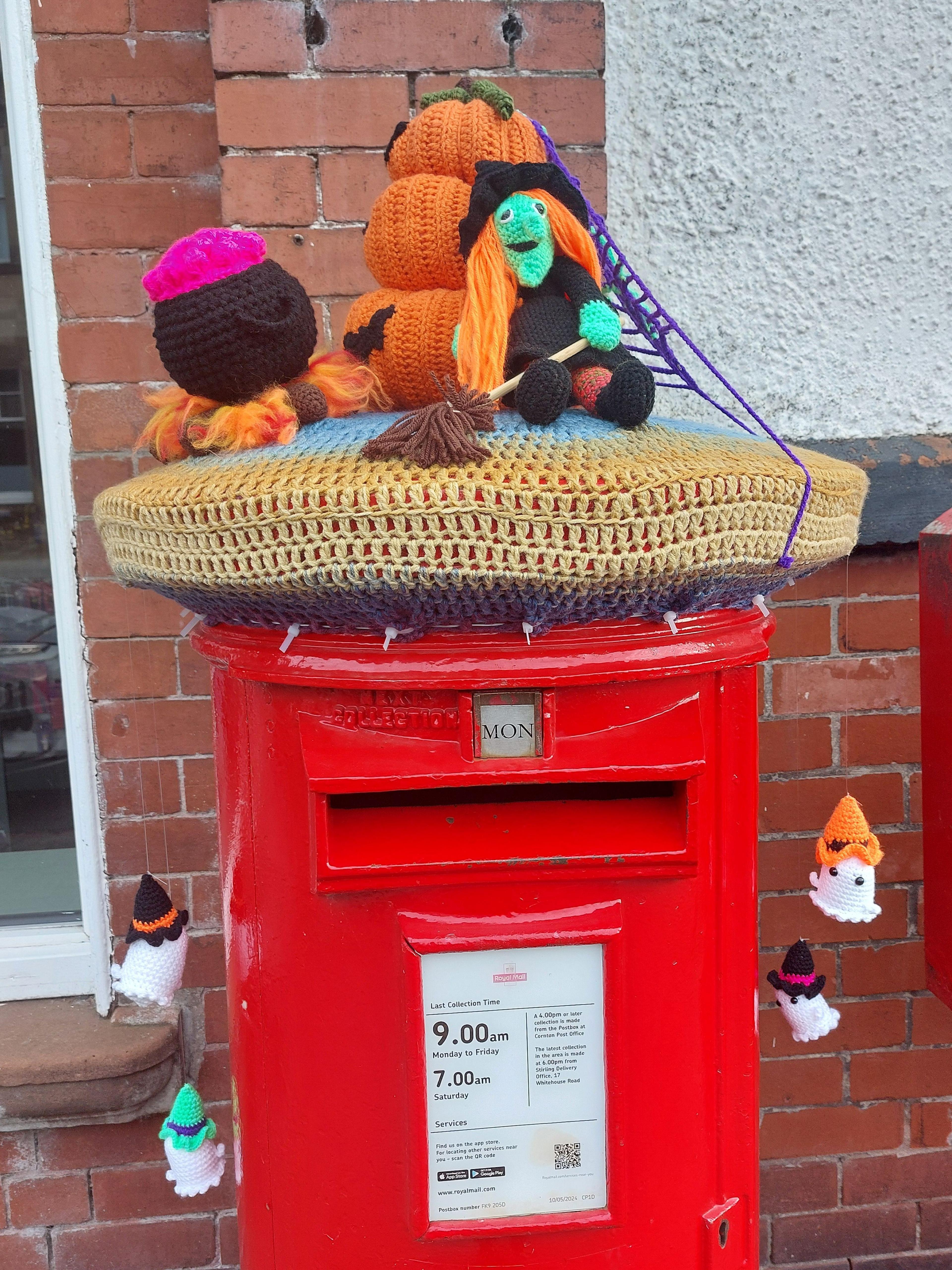A post box with decorations for Halloween - including a knitted witch and knitted pumpkins placed on top