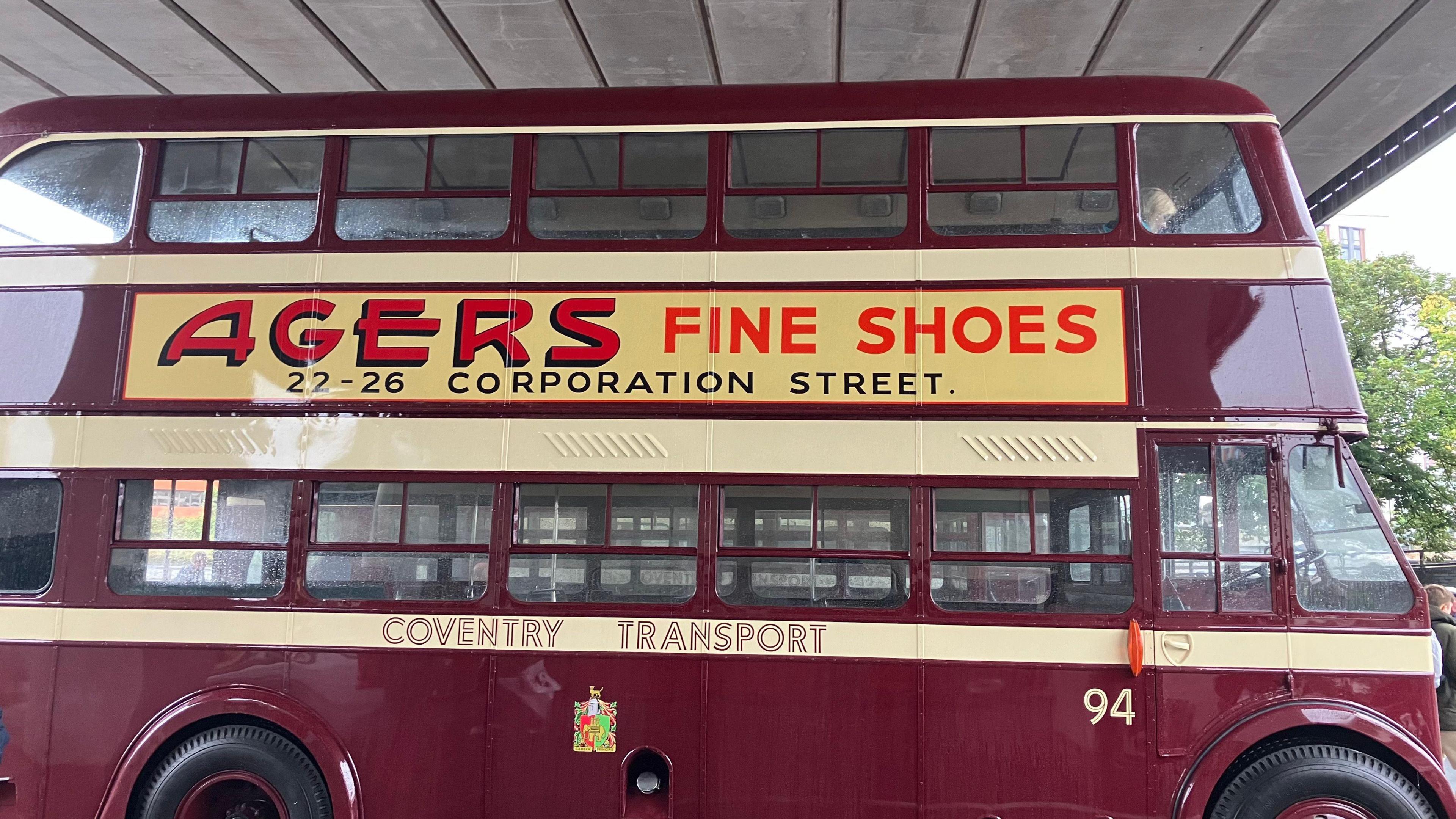 A side-view of a vintage red double-decker bus, with a pale yellow advertising sign which says 'Agers Fine Shoes' in a bold red print and '22-26 Corporation Street' underneath in black. Painted on the side of the bus are the words 'Coventry Transport'.