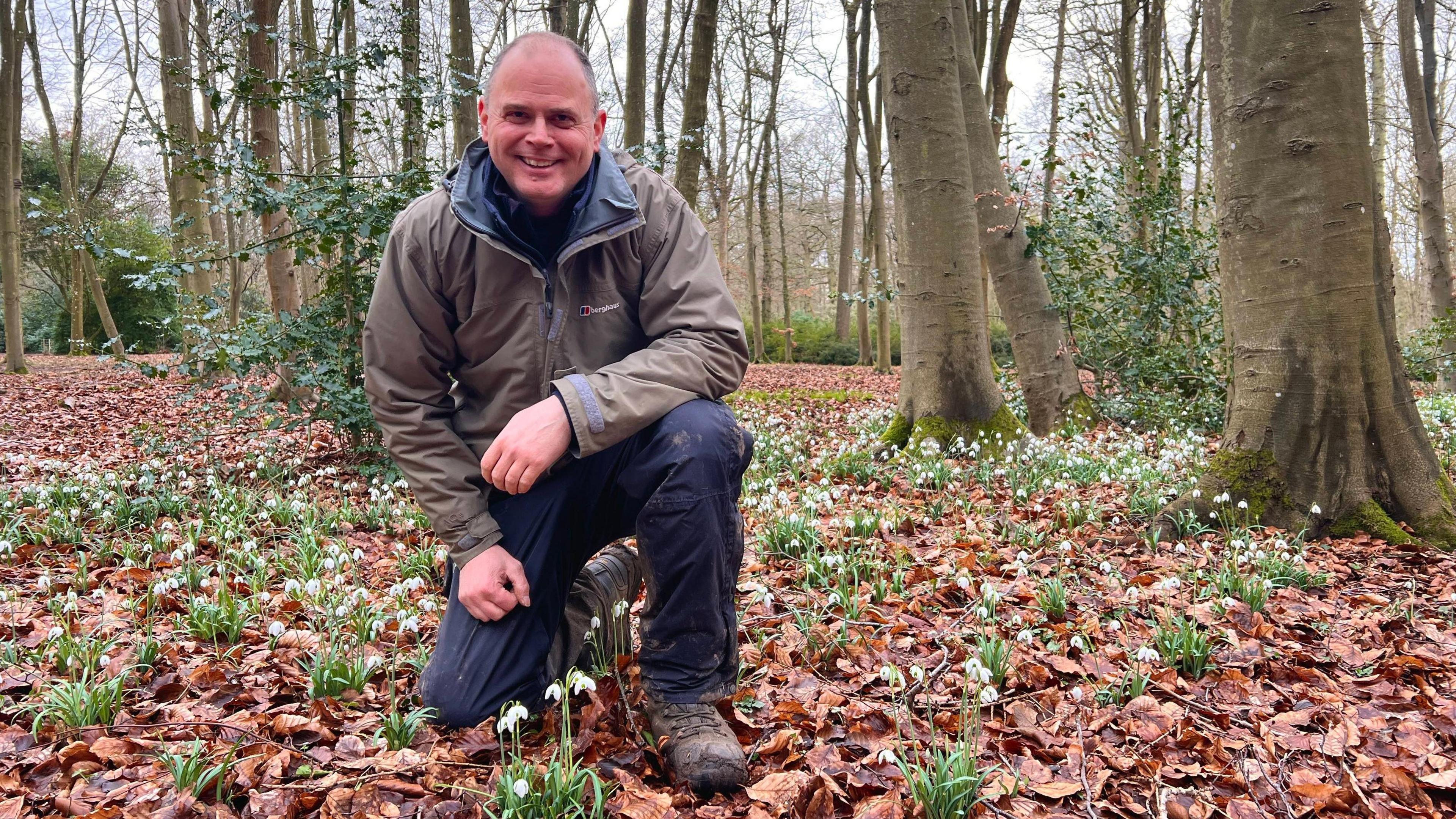 A smiling Dan Winter, the head gardener at Evenley Wood Garden, kneels down on the forest floor amidst snowdrops that have pushed their way up through soil and dry leaves 