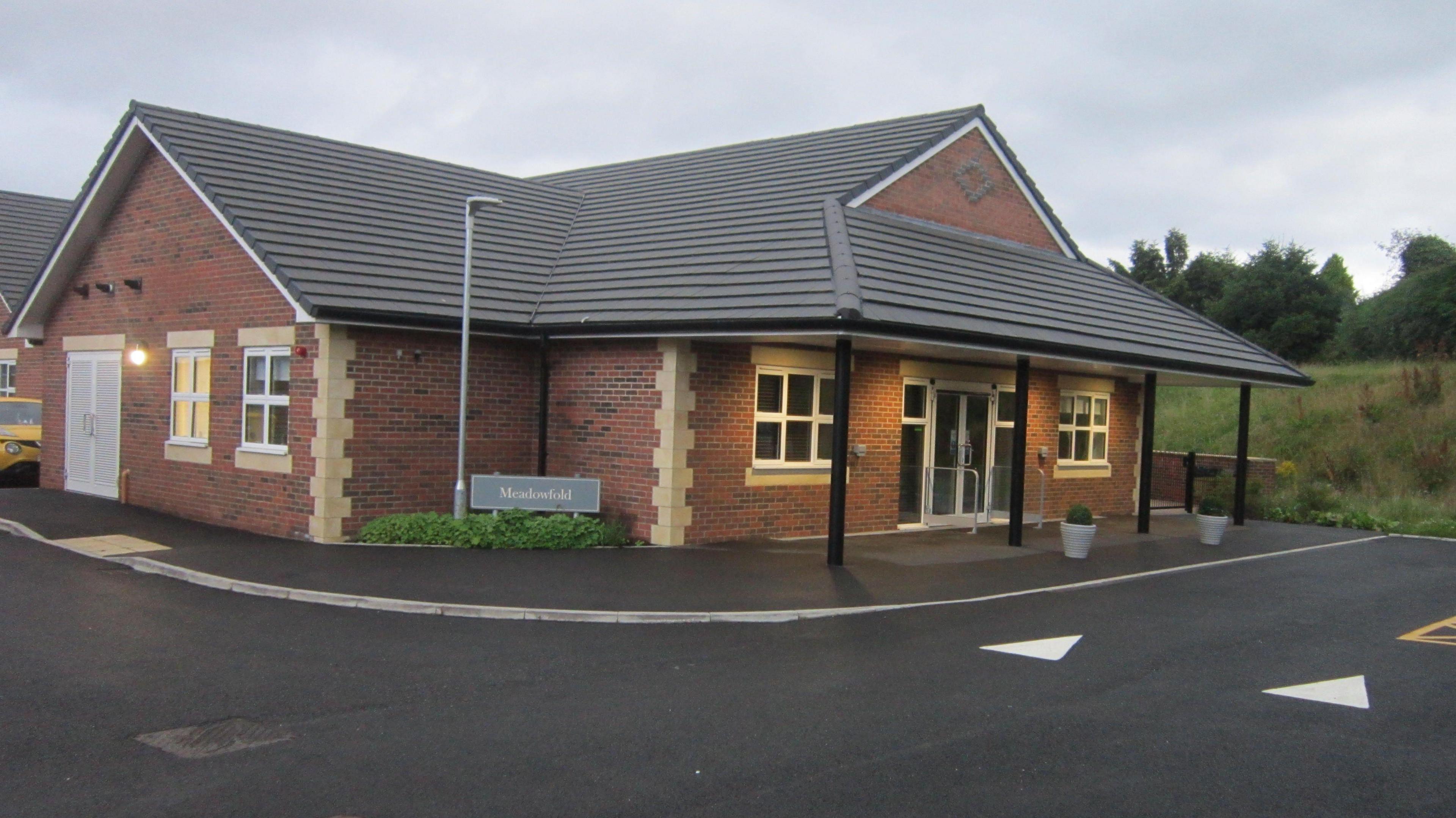 A view of the red brick building with a tarmac driveway in front and trees and grass to the rear