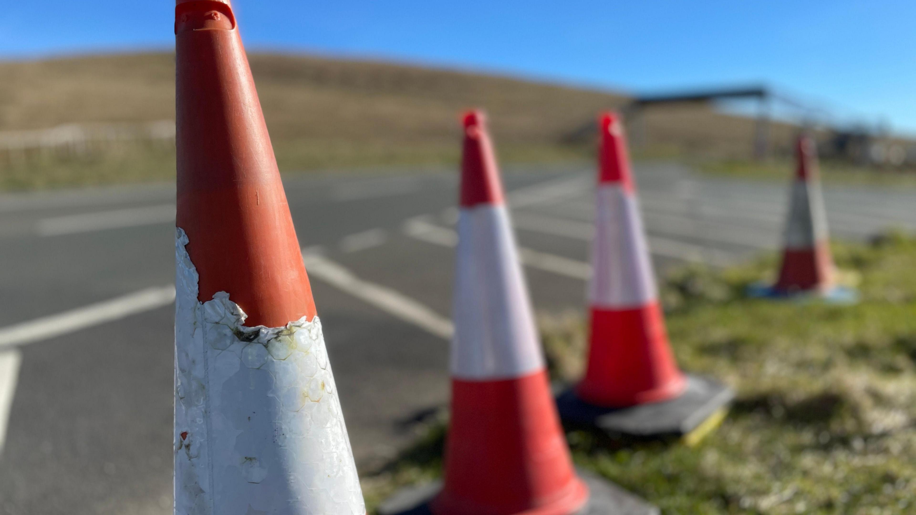 Orange and white traffic cones on the side on the Mountain Road, with a metal footbridge over the carriageway in the distance.