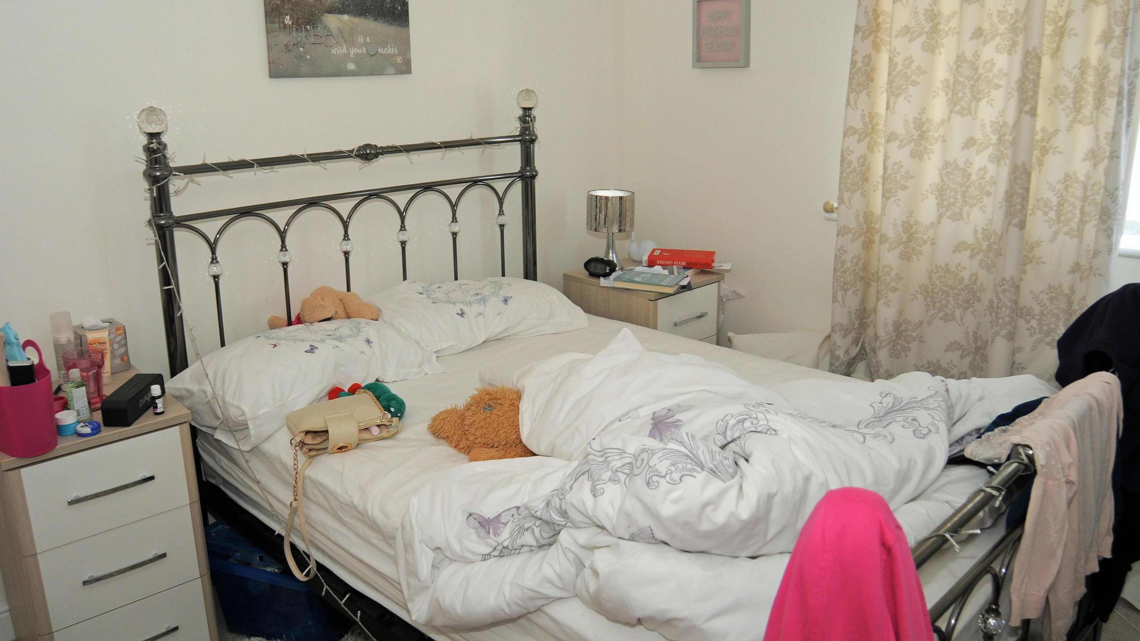 An unmade bed with a cream coloured handbag and a teddy bear on the mattress, which is covered by a white sheet. 