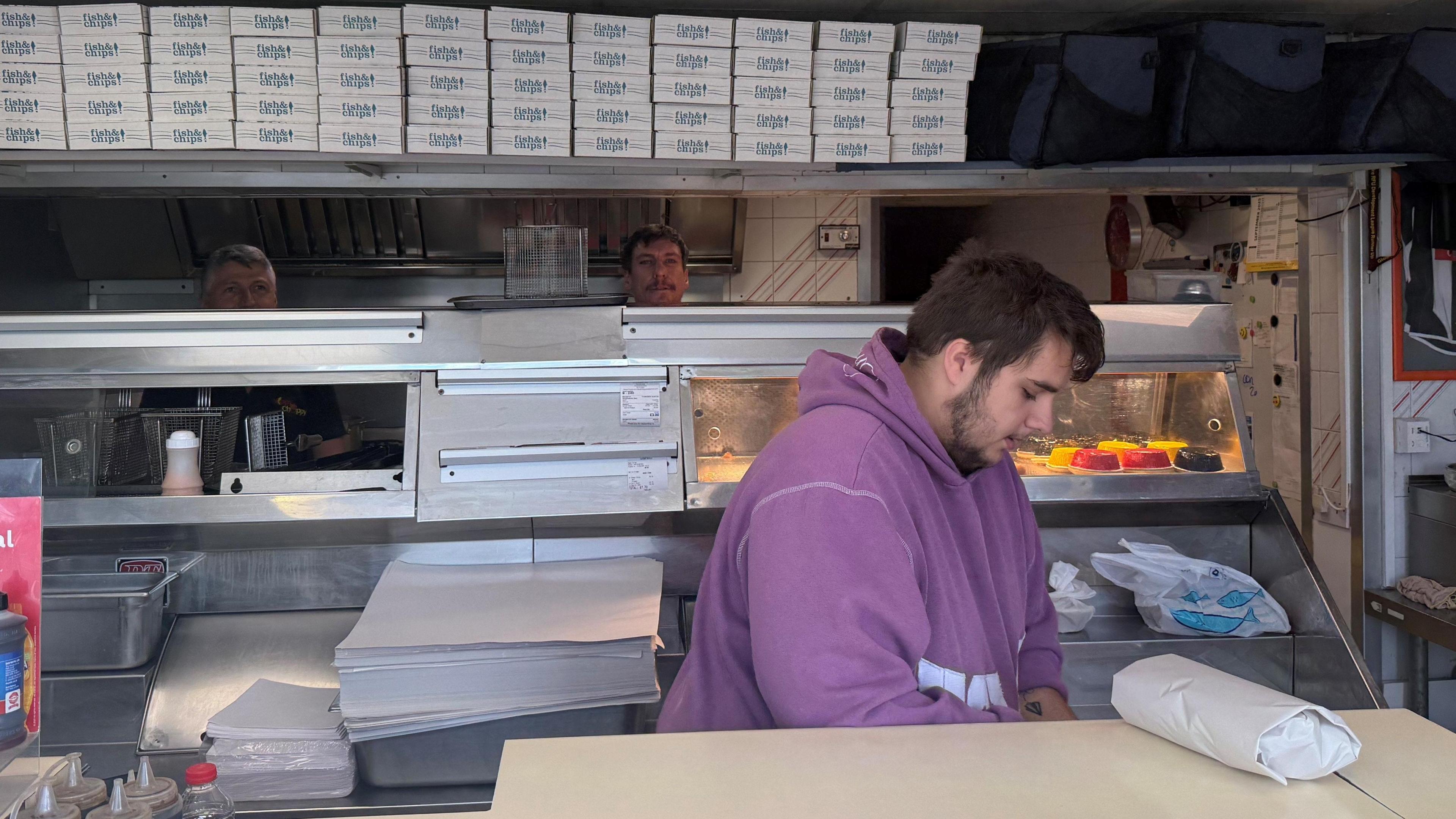 A man working behind the counter at the Nippy Chippy in Stonehouse, Gloucestershire. He is wearing a purple hoodie and looking down. 
There is a parcelled portion on the counter and empty boxes behind him with Nippy Chippy on them. 
There are two other members of staff in the background at work. 
And a warm plate between the front counter and the staff at the back.