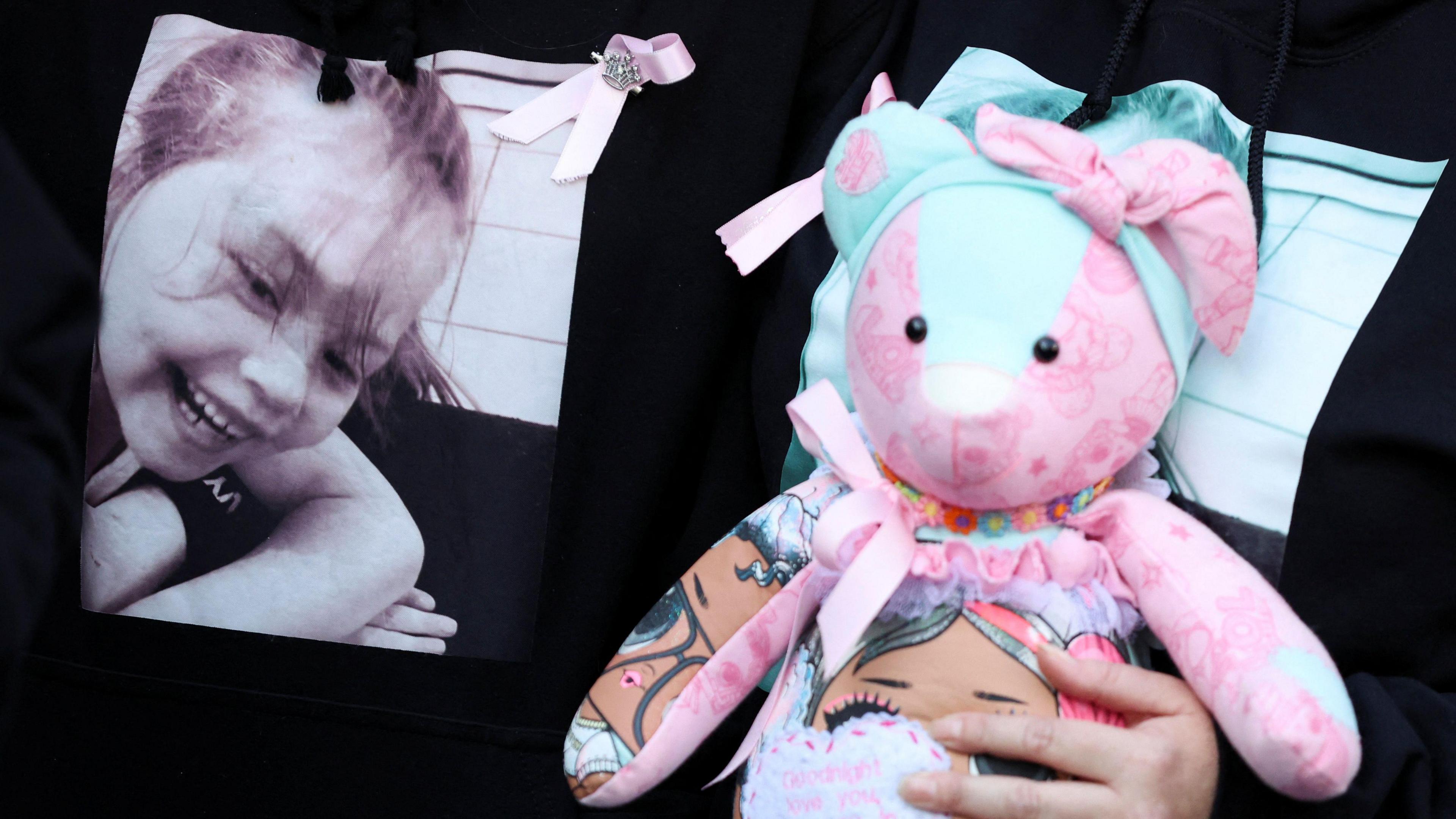 A woman holds a pink and light blue teddy bear to her chest, covering a T-shirt with a black and white picture of Olivia Pratt-Korbel smiling on it. Another woman wear the same T-shirt alongside her. Only their torsos are in shot
