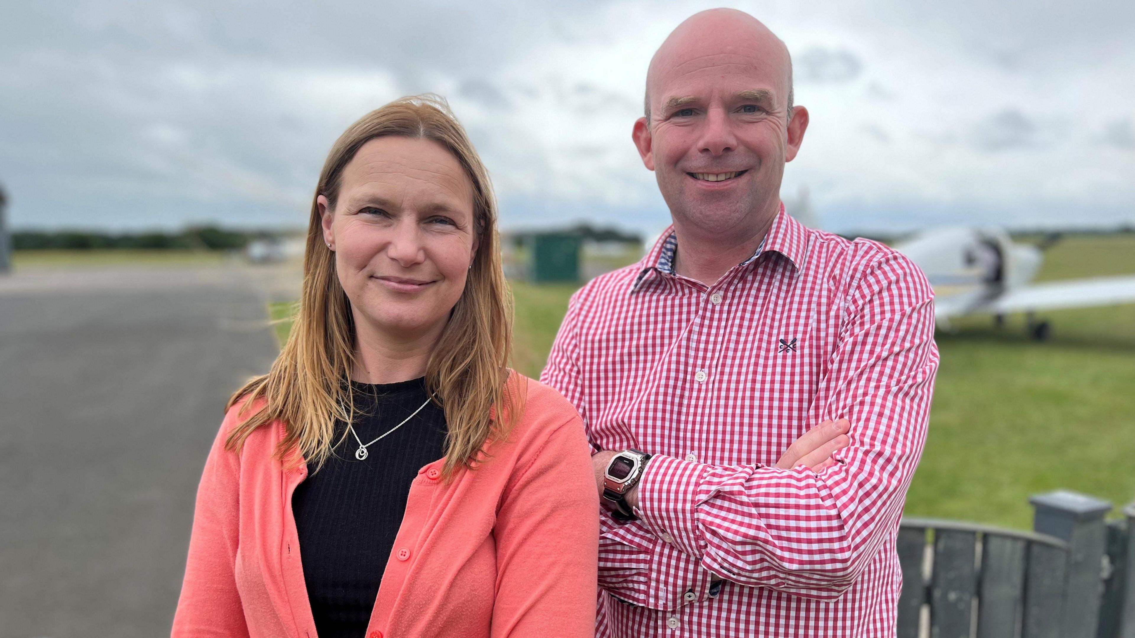 A woman and a man stand next to each other at an airfield, with a white light aircraft on a green field in the background. The woman has long light-brown hair and wears a black top, pink cardigan and silver necklace; the man has a shaven head and wears a pink and white check shirt and a black and silver watch.