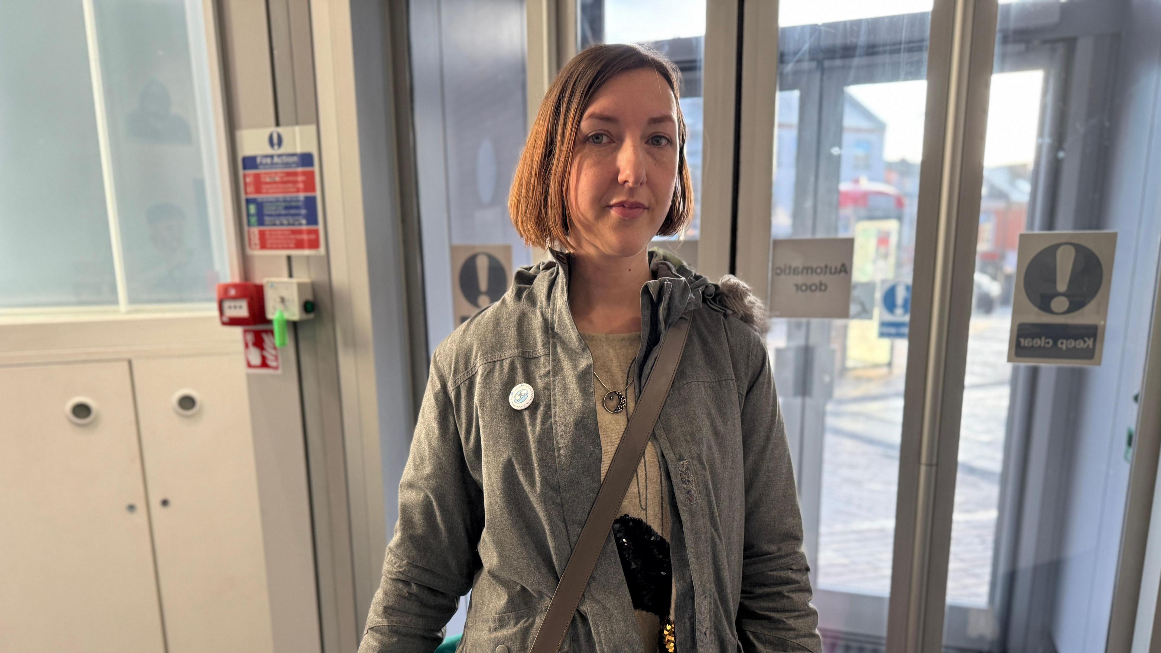 A woman in a grey coat with a brown bag over her left shoulder poses by some glass doors.