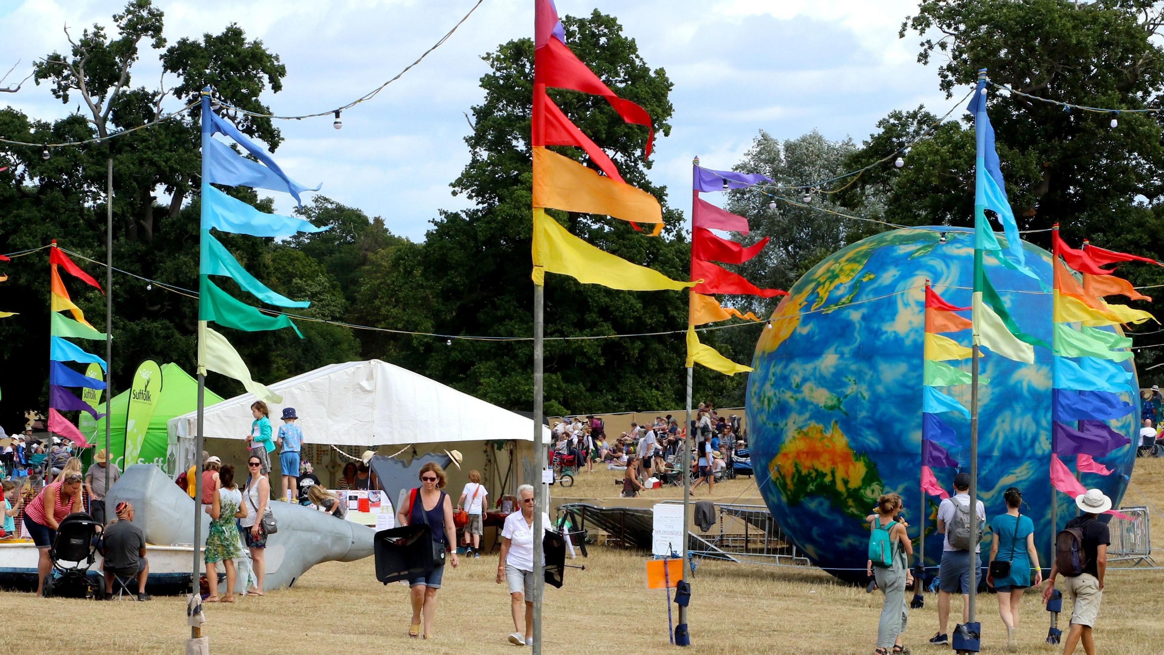 People wander about a festival. Multi-coloured festival flags have been erected and can be seen blowing in the wind. Tents and festival structures can be seen dotted about.