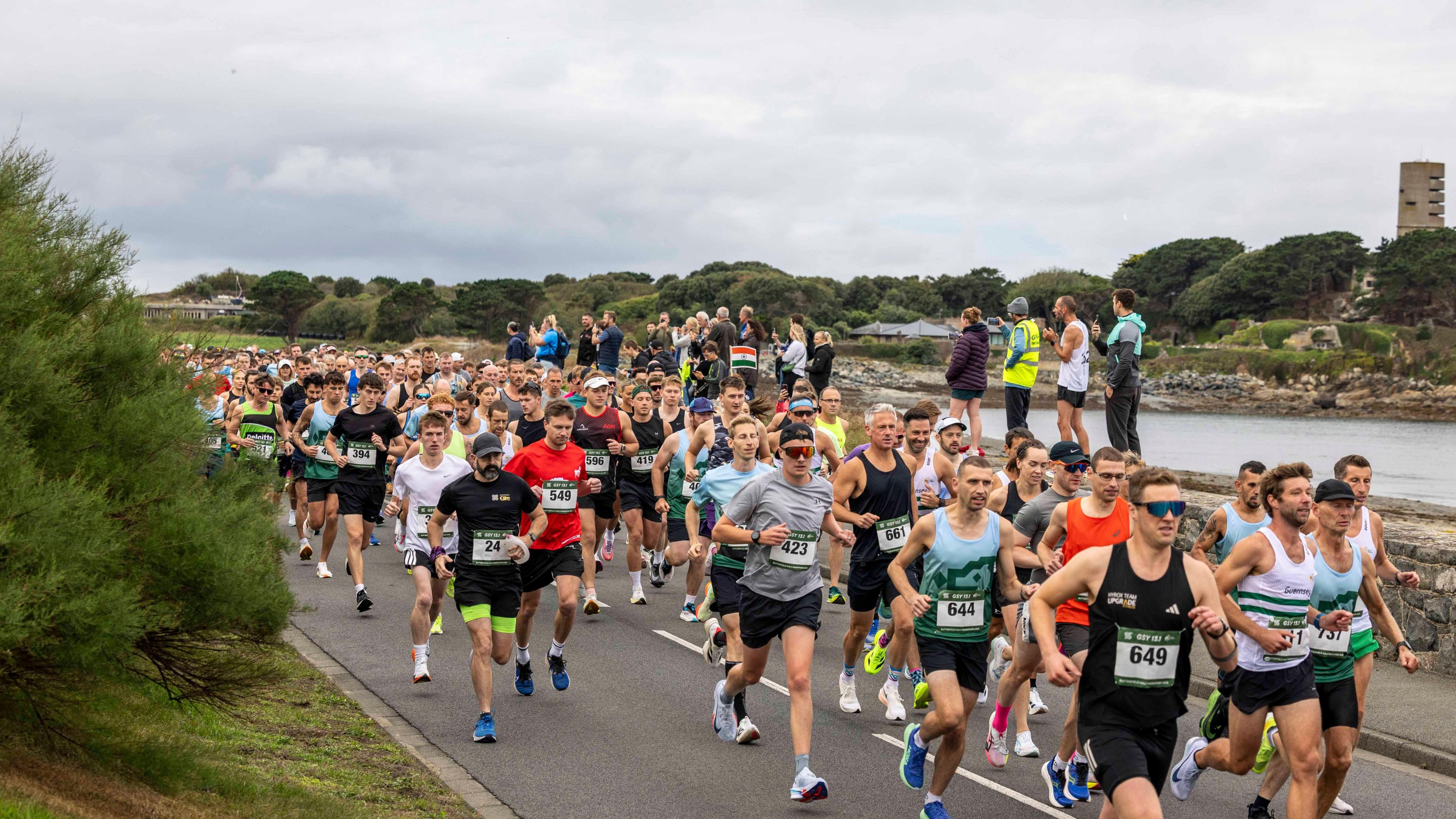 Hundreds of people set off on a half marathon, all dressed in running gear with numbers on their chests. Spectators watch with a small cove and World War Two concrete gun emplacement behind them,