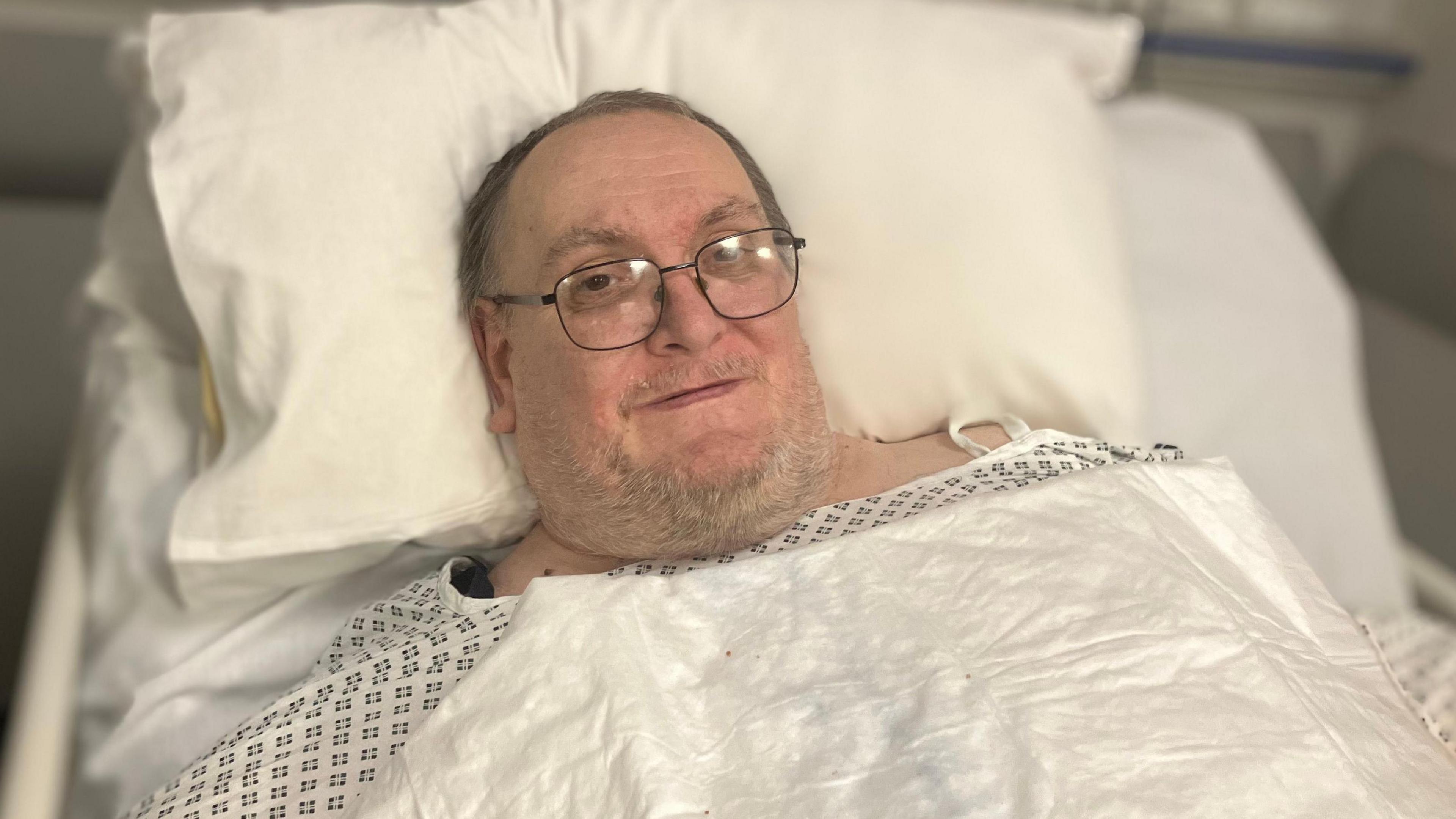 Simon Morris has grey hair and glasses and  is wearing a white hospital gown and is lying in a  hospital bed.  