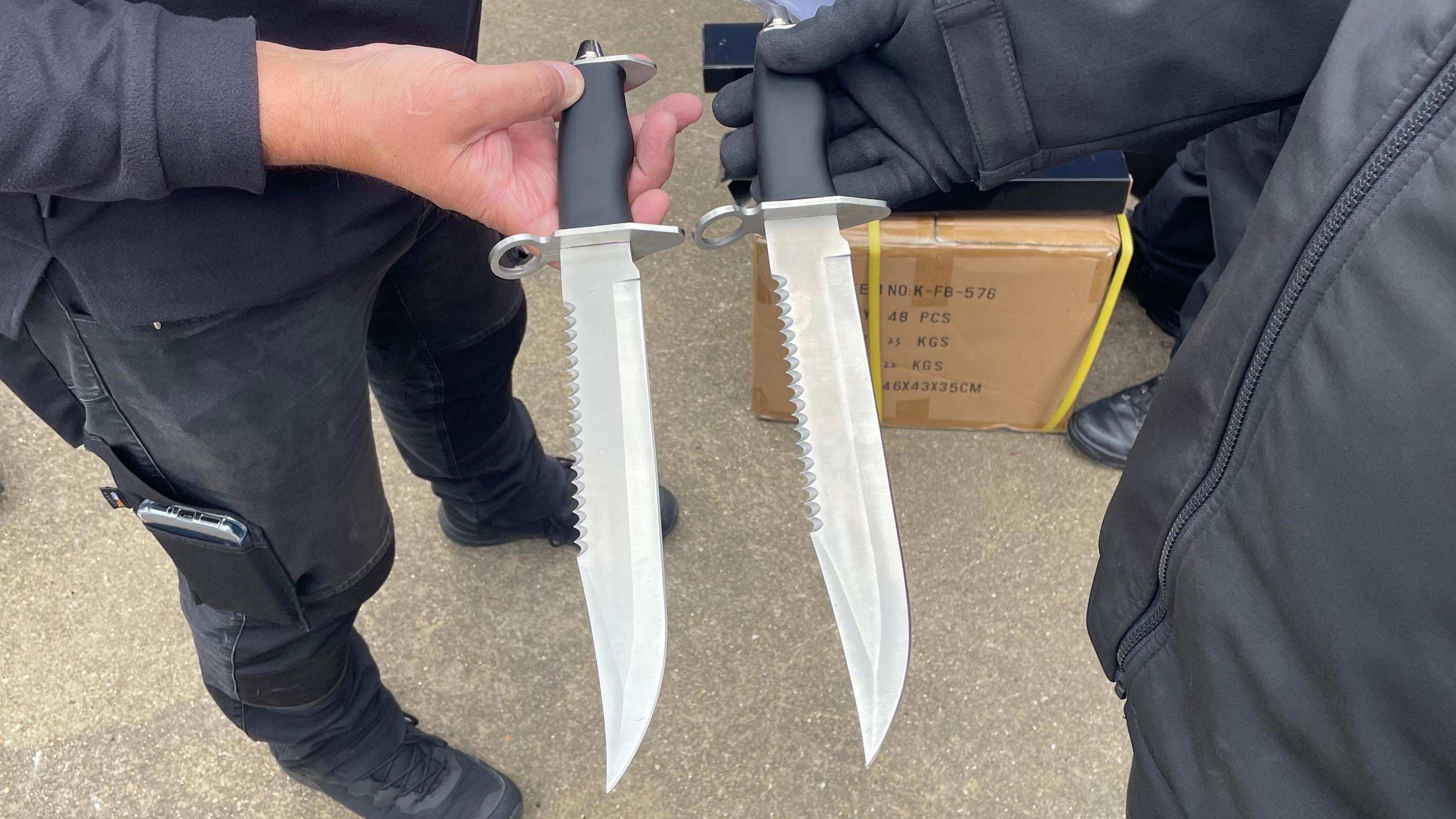 Two seemingly identical knives held by two police officers. The knives have sharpened blades, as well as lengths of serrations along the top of the blade. Their handles are black and both have circular metal fixings, apparently so they could be used as bayonets. They are held with their points facing towards the bottom of the frame. The concrete ground can be seen in the background along with a cardboard box and one of the officer's legs and feet.