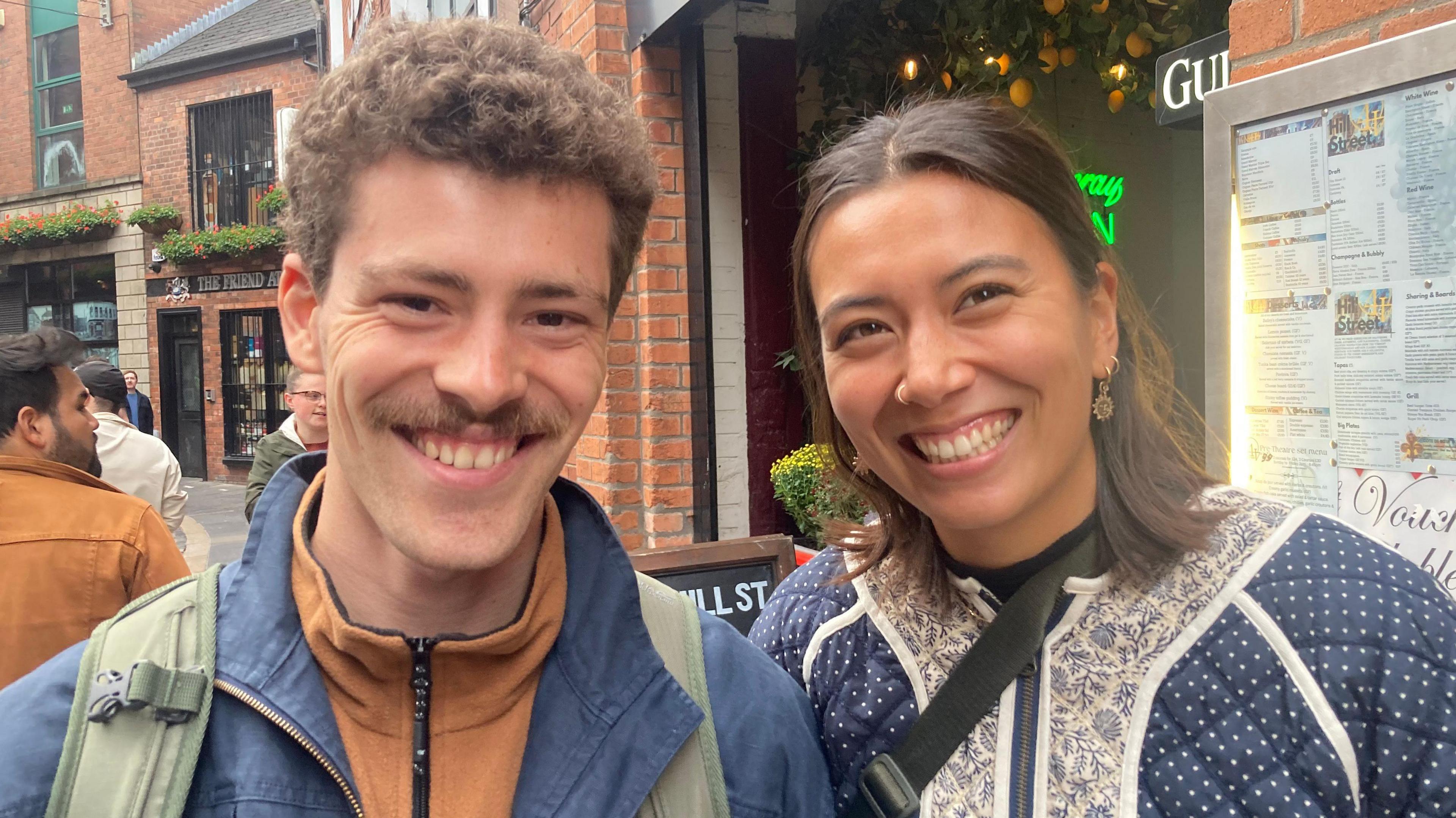 A Canadian couple called Tibo and Dayna standing in a street in Belfast city centre, smiling at the camera.  Tibo has short, curly fair hair and a moustache and is wearing a brown zipped sports top and a blue jacket. Dayna has shoulder-length straight brown hair and is wearing a padded navy jacket with white polka dots and a white lace collar. They are both carrying shoulder bags. 