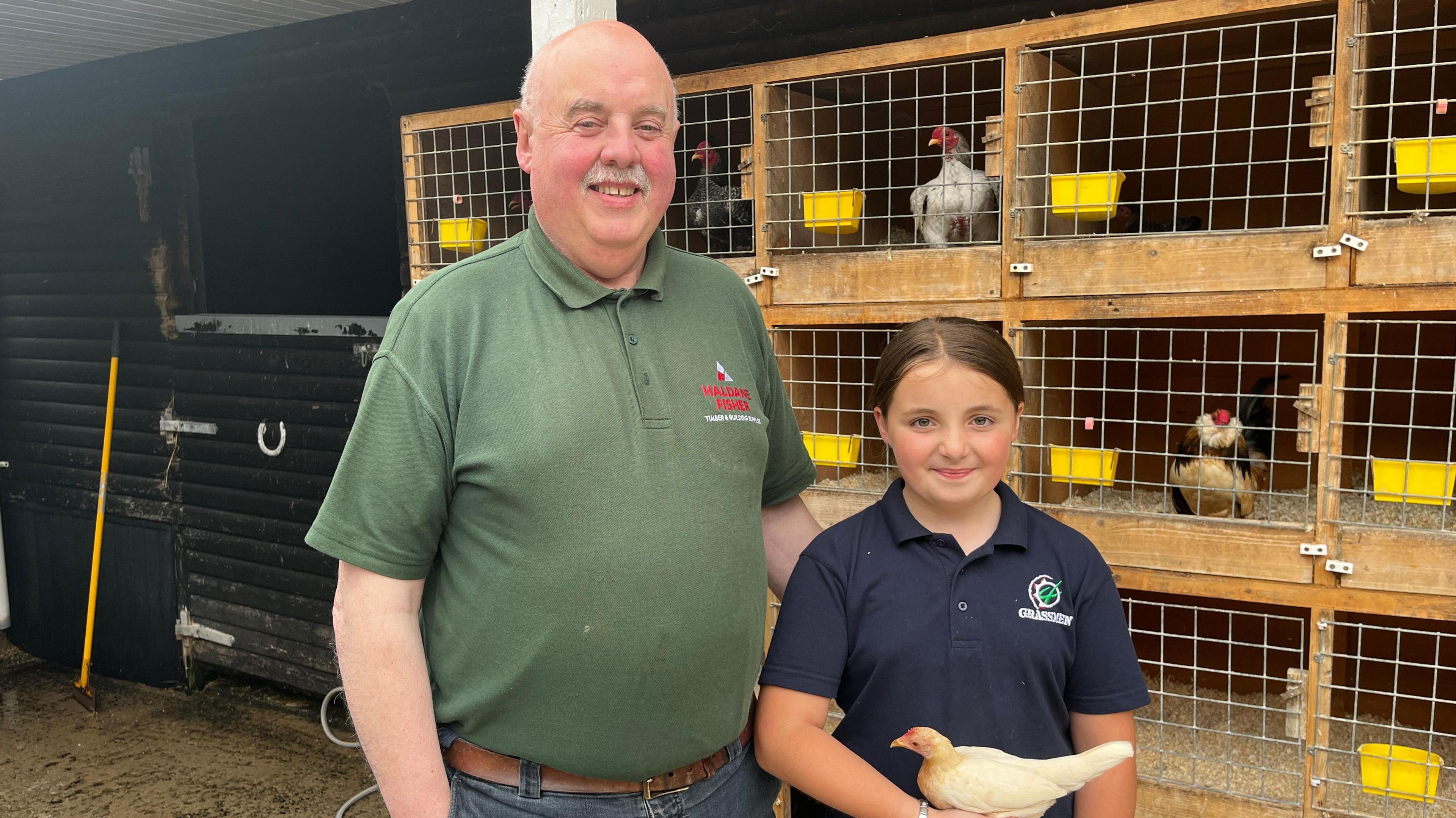 Alannah (right) stands next to her granda Joe (left). He is wearing a green polo shirt and has his hand on Alannah's back. Alannah is wearing a navy polo shirt and is holding a small chicken. In the background, several chickens are in enclosures.