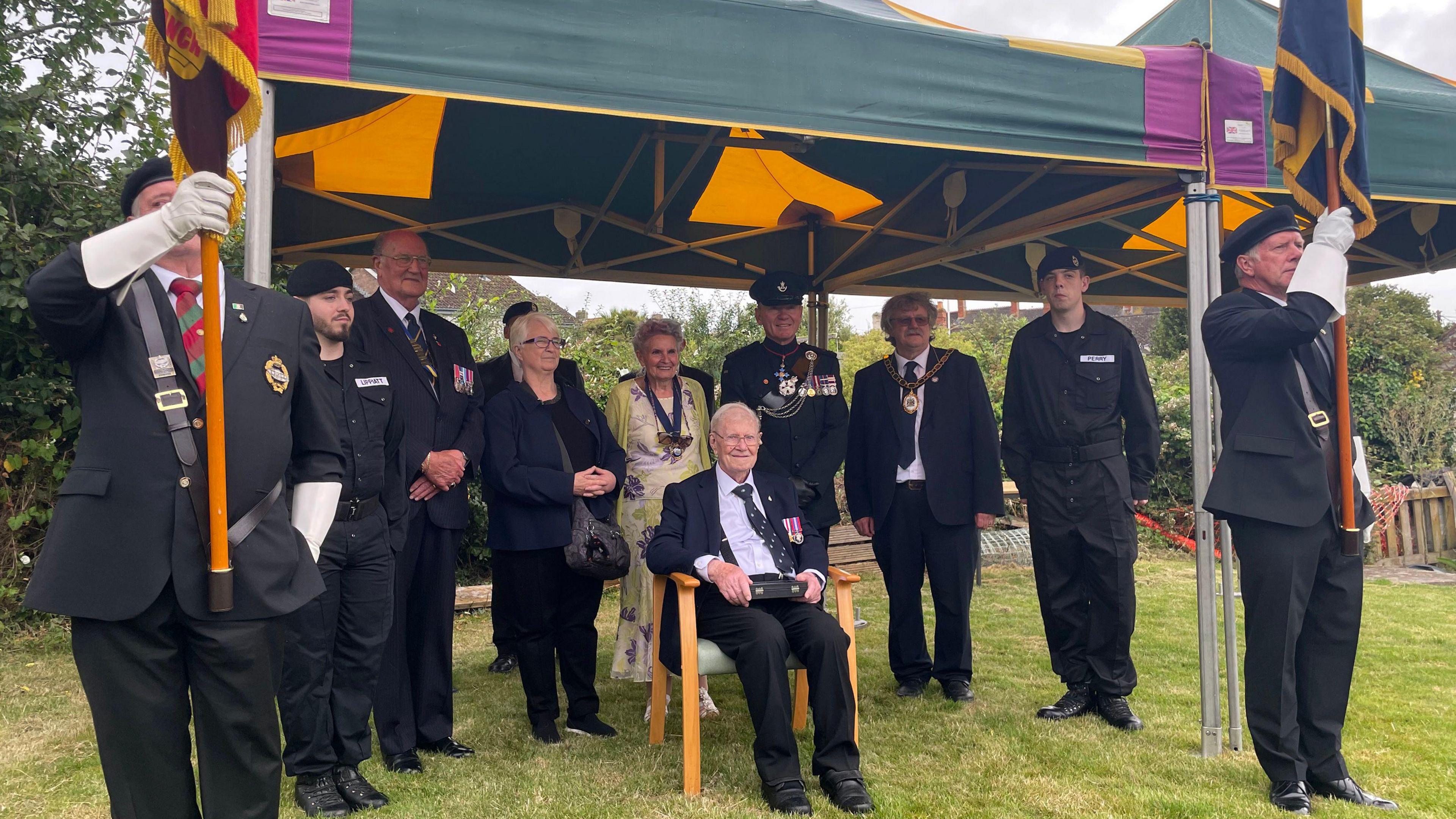 The ceremony during which George was presented with his medals. He sits on a wooden frame under a gazebo, with two men in military dress holding flags. A number of other members of the Royal British Legion and his family stand under the gazebo. 