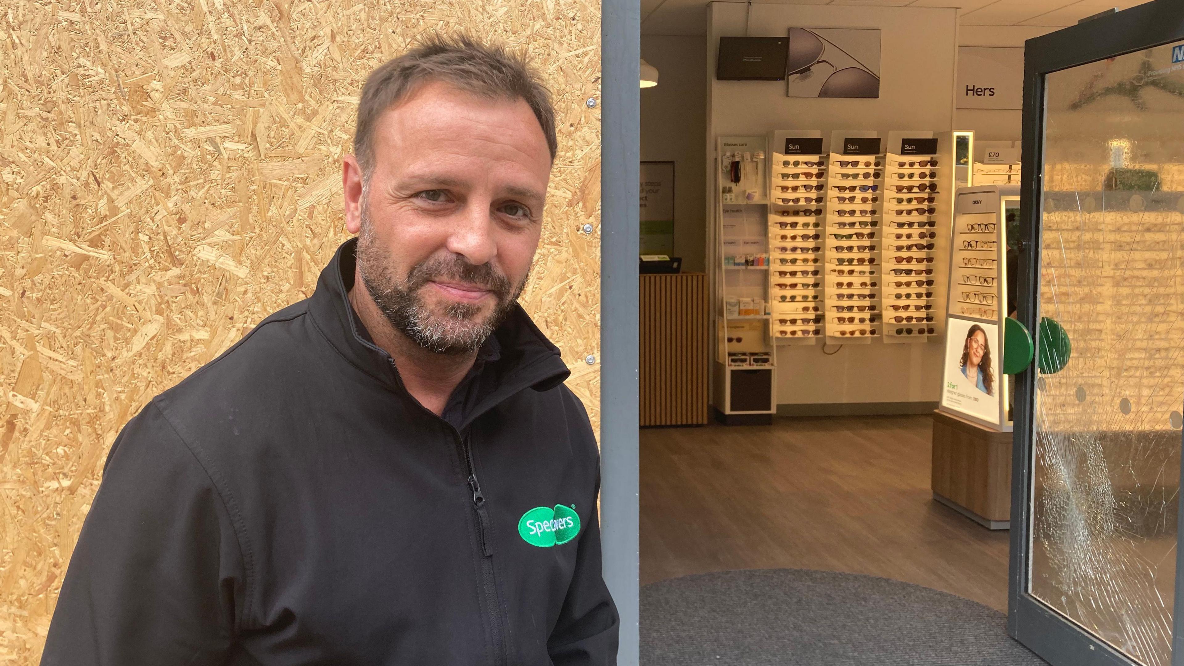 David Proudfoot is wearing a black jumper with a green Specsavers logo on it. The window behind him has been boarded up and another is badly smashed