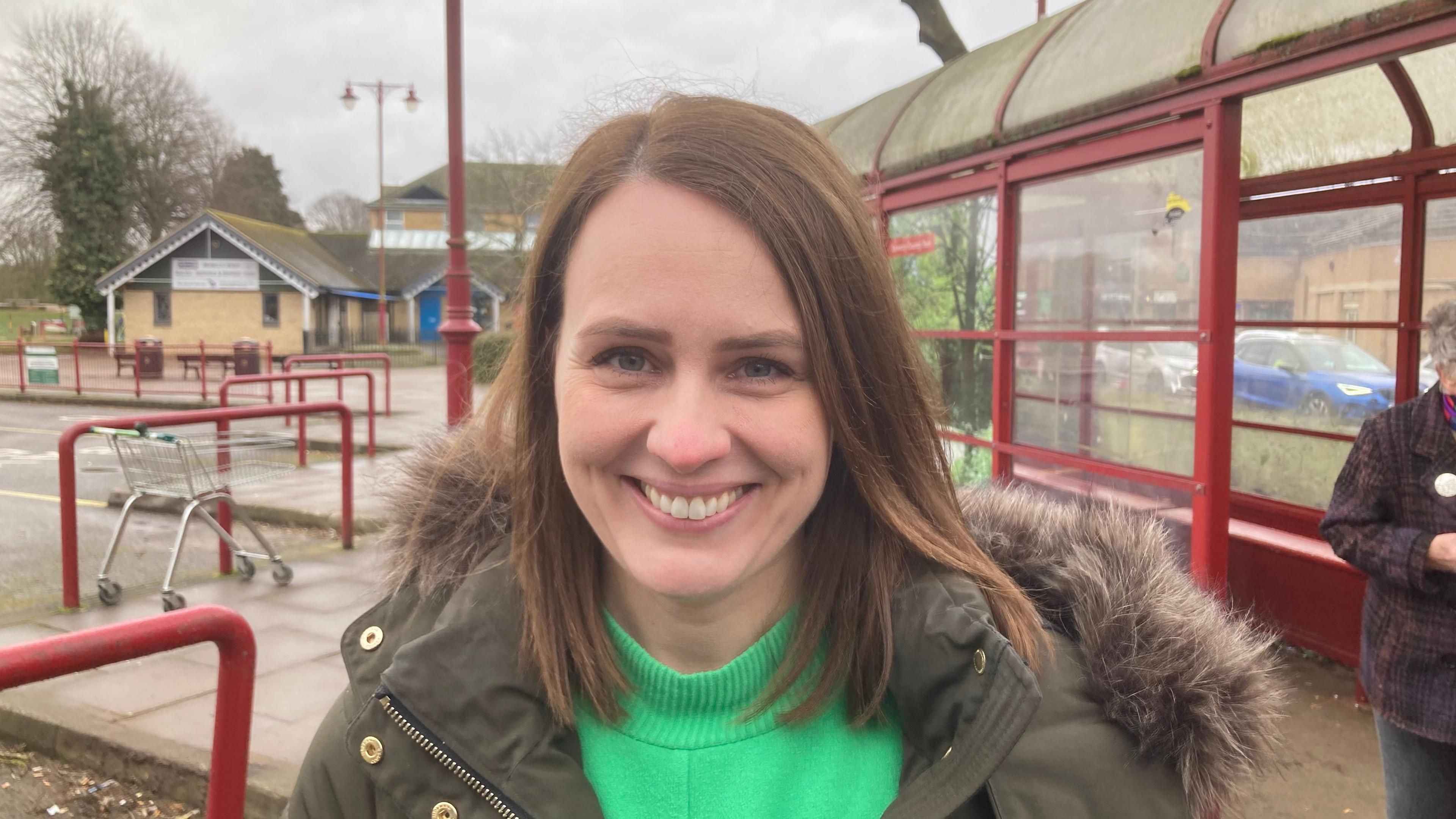 Hayley Charlesworth with long brown hair smiling at the camera. She is wearing a green coat with a furry hood and a light green top. There is a red-framed glass bus shelter behind her. There is a shopping trolley to the left.