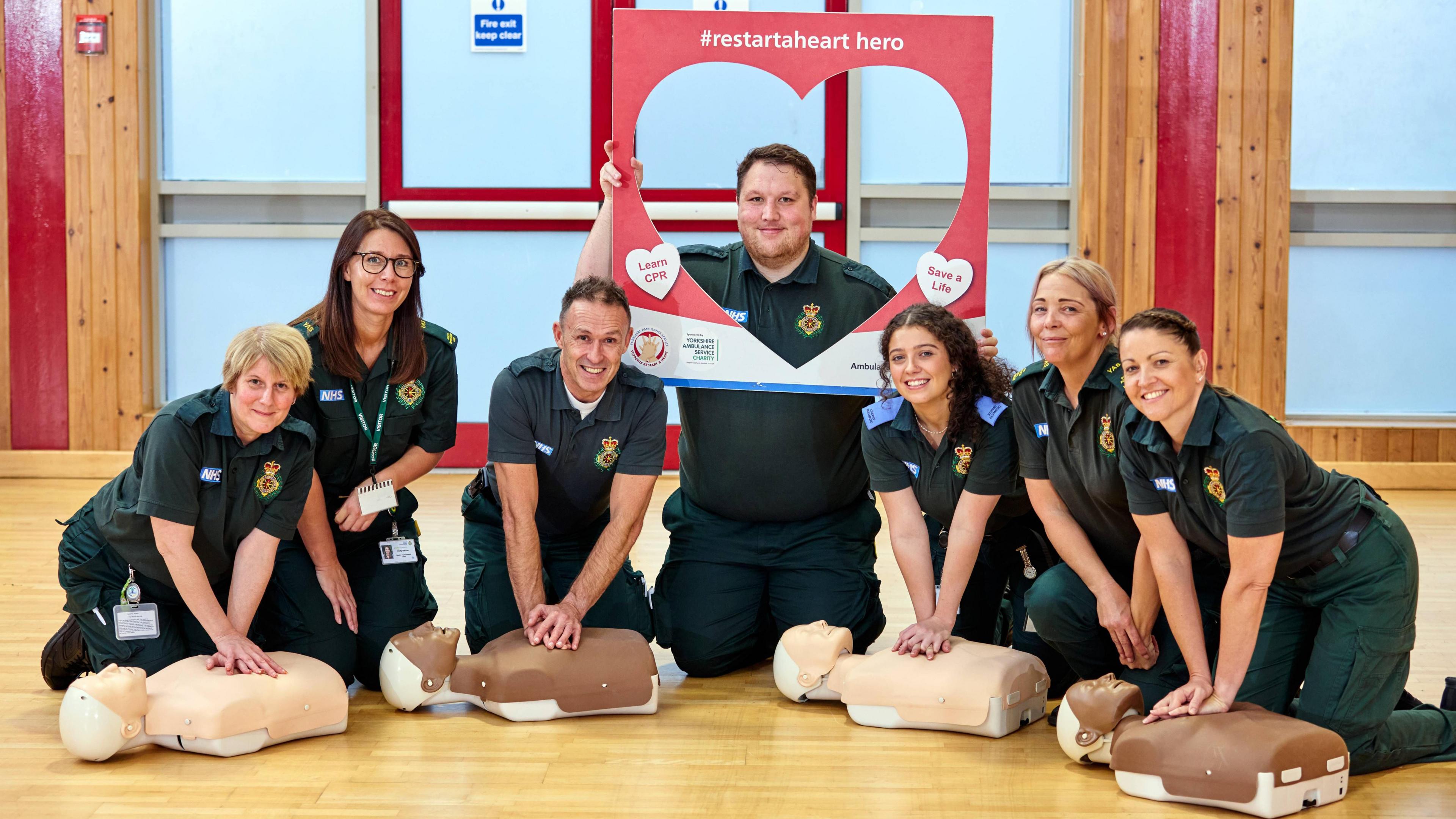 Seven Yorkshire Ambulance service staff pose on their knees - four with mannequins in front of them, and one holding a cardboard cut-out saying restartaheart hero, Learn CPR, Save a Life on it. They are all in their dark green paramedic uniforms on a wooden floor.