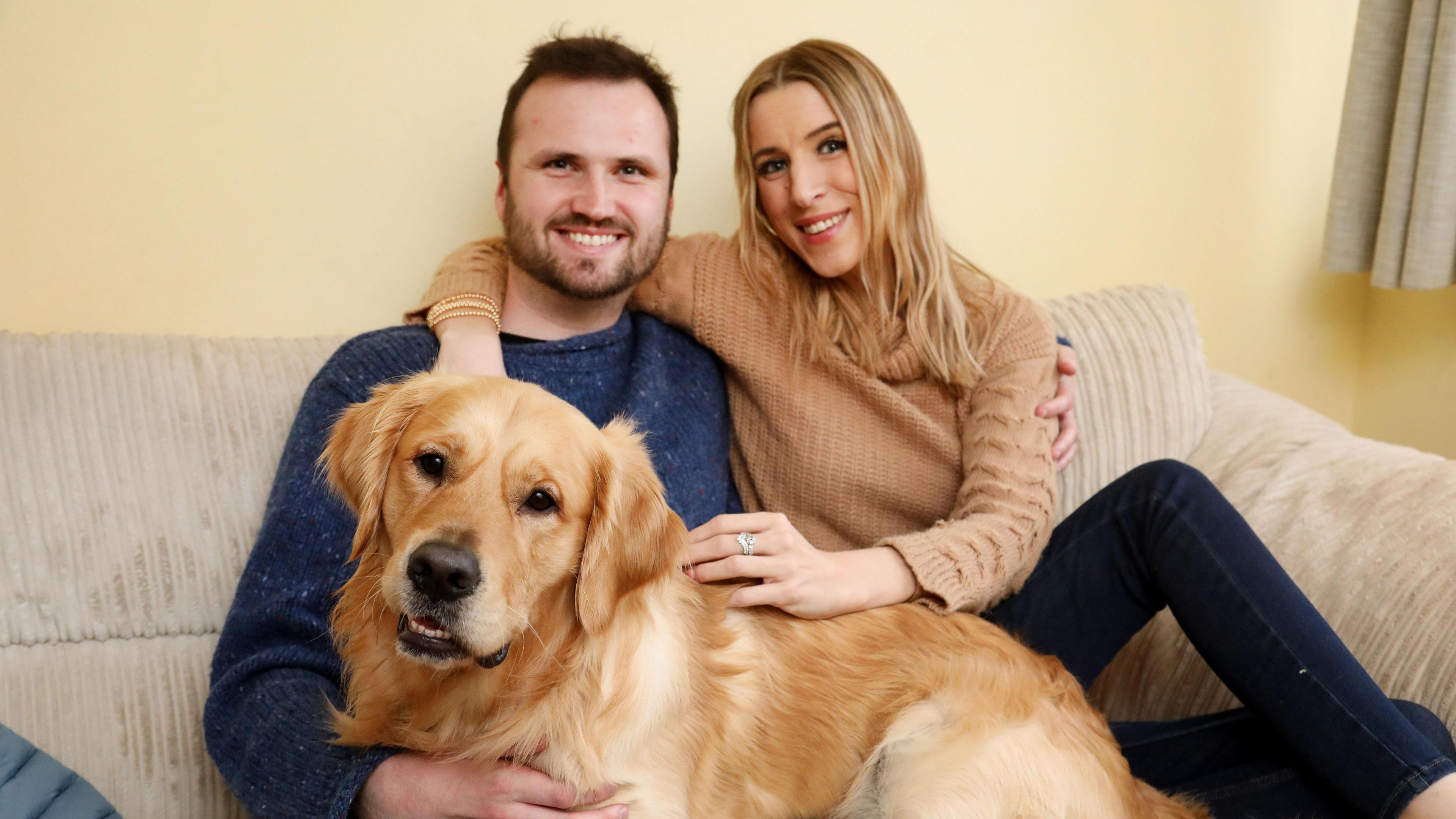 Faye Louise sat with partner Will and dog Neville. They are all looking at the camera. Faye and Will are smiling and have their hands around one another. 