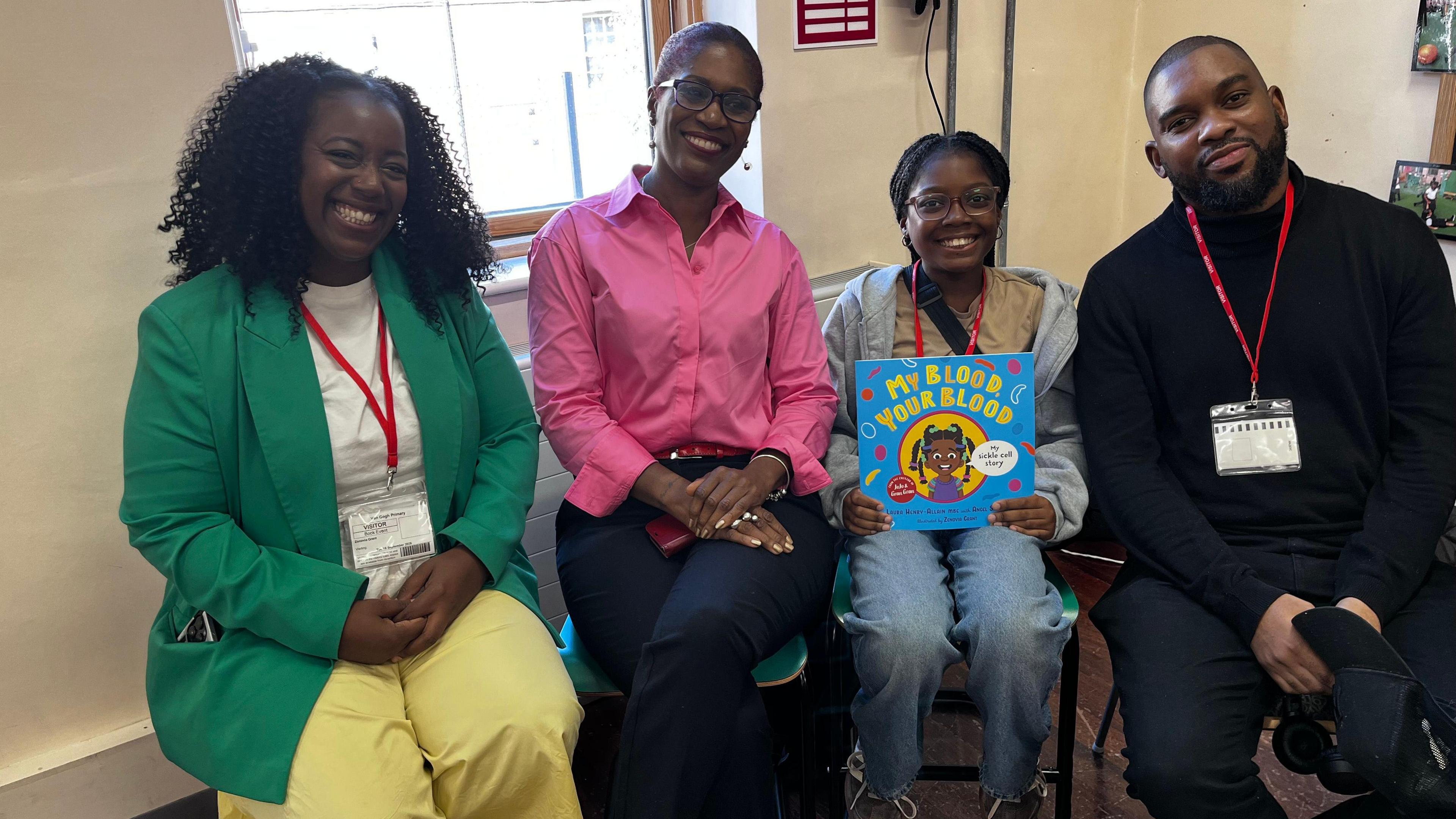 Four people sat on chairs in a room in a classroom, with a window and white walls behind them. All four of them have dark skin and are smiling at the camera. The woman on the far left is wearing a green blazer and yellow trousers. The woman to her right is wearing a pink shirt and dark blue trousers. Angel and Kehinde are sat on the right, with Angel holding a copy of the book, 'My Blood, Your Blood'.