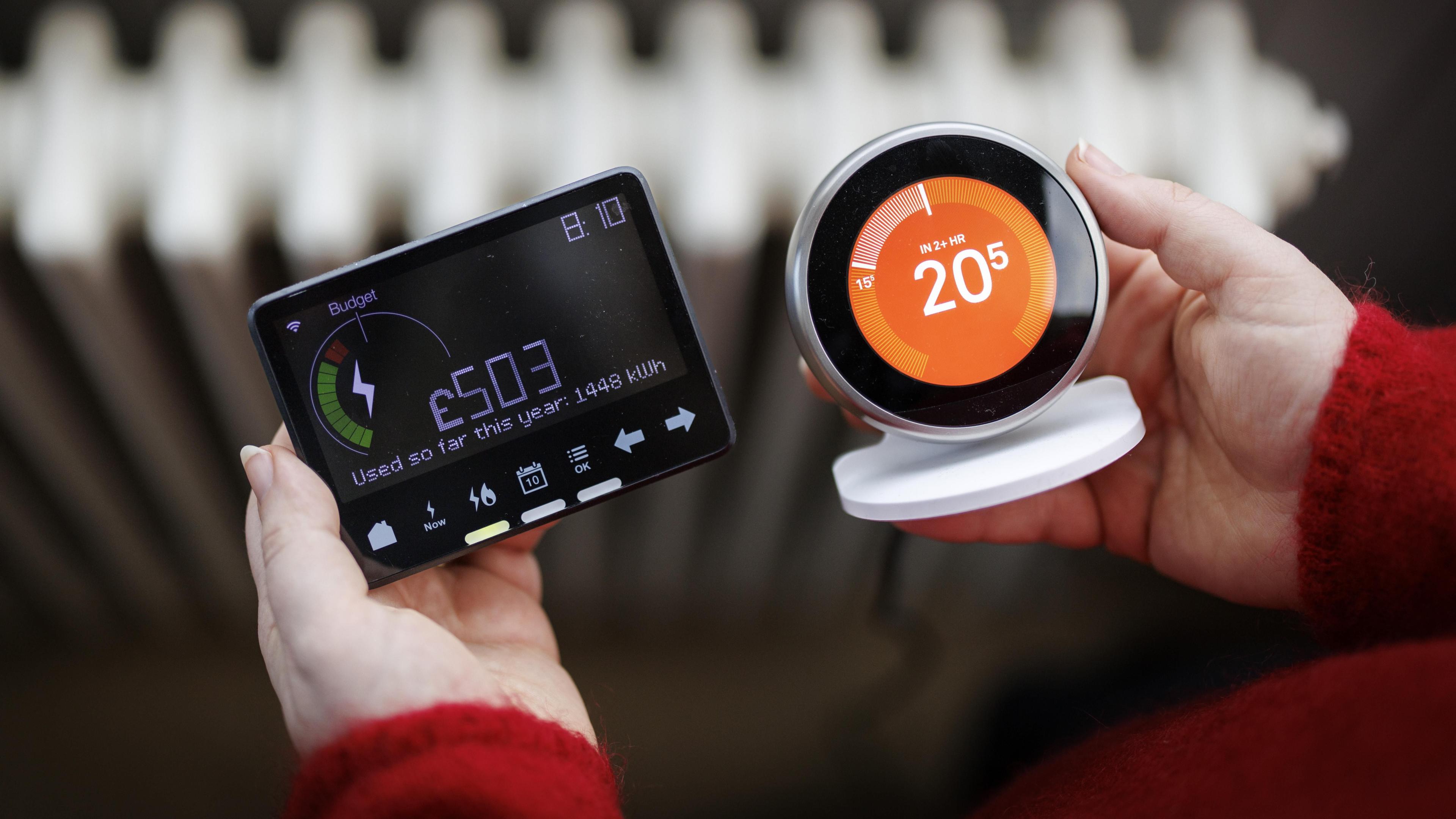 A person holds a smart energy meter, used to monitor gas and electricity use, with a smart thermostat in a home in London