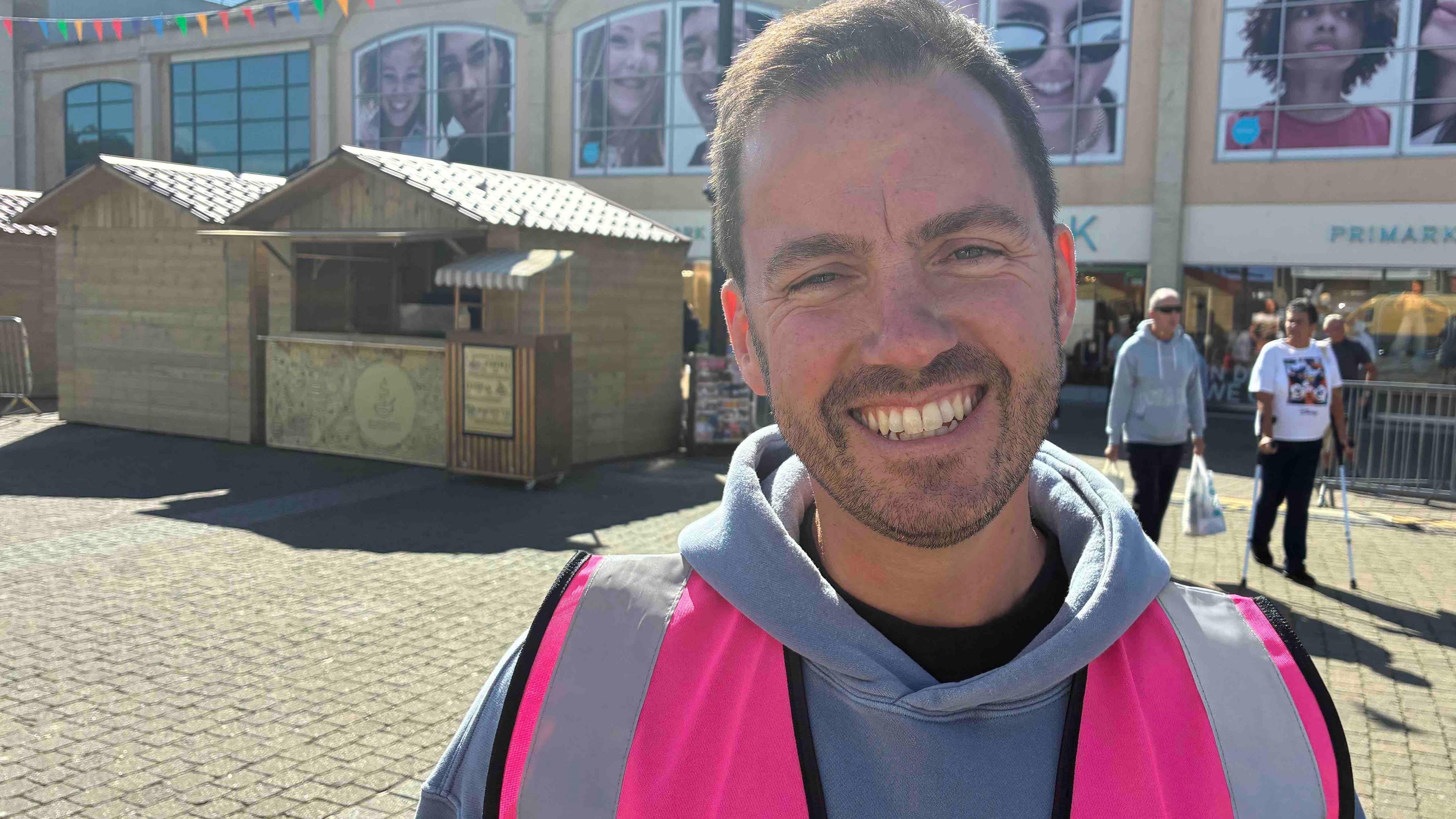 A man in a pink high vis jacket is smiling on a sunny day in front of wooden market stalls.