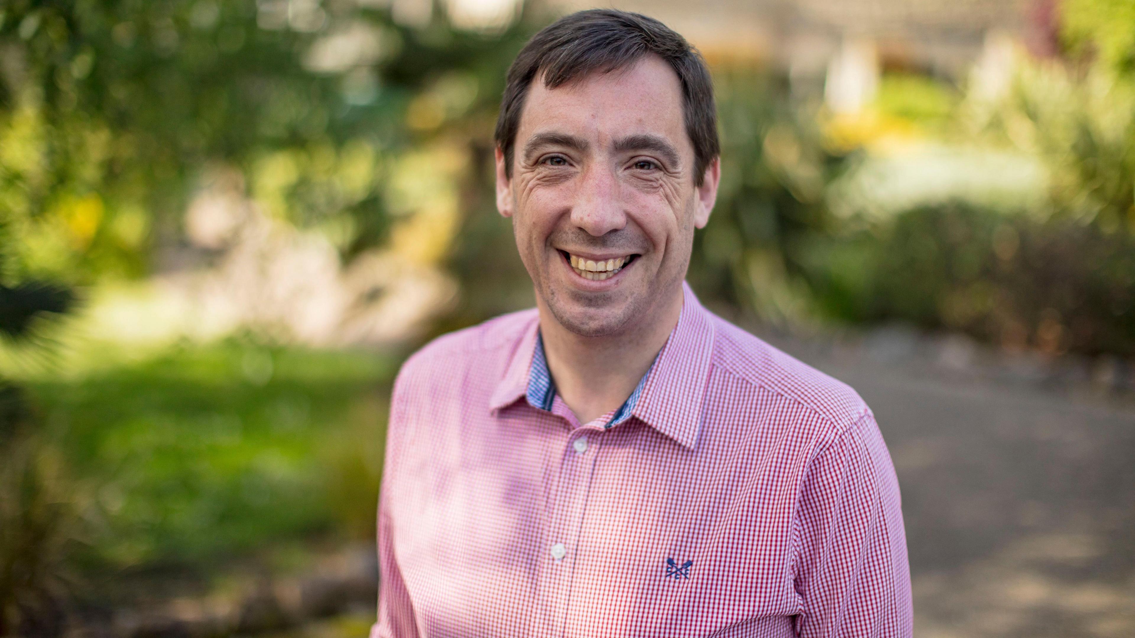Alan Williams. He has short dark hair and stubble on his chin. He is wearing a red and white checked collared shirt. He is looking straight ahead and smiling