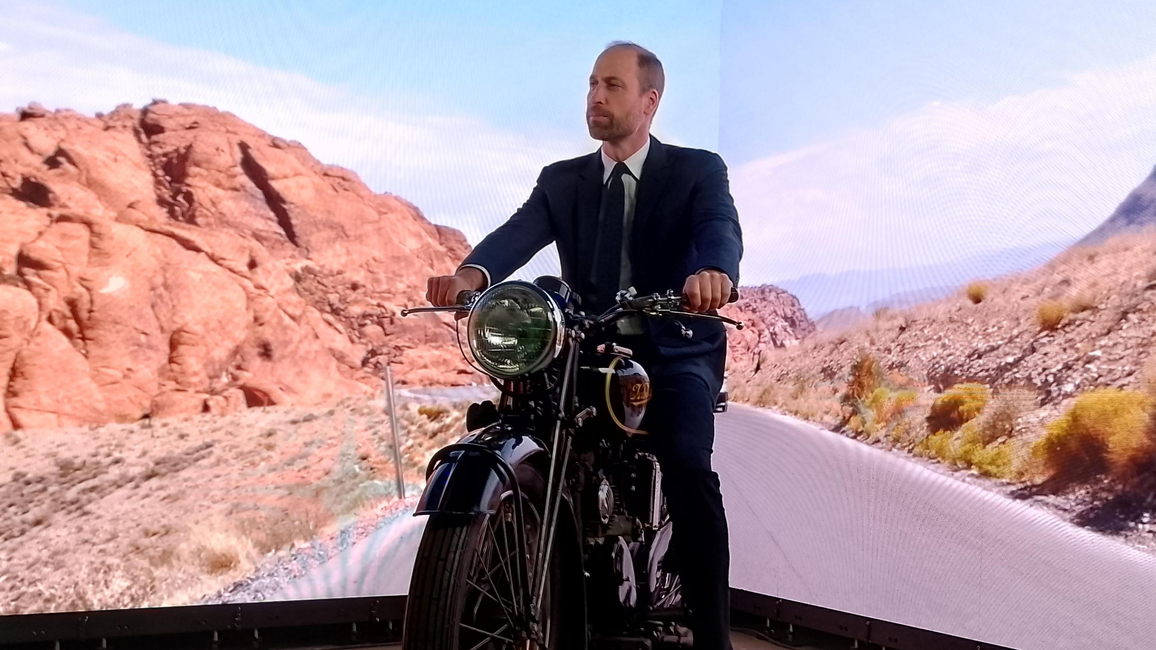 Prince of Wales on a motorbike in front of a green screen showing a road and mountains. 