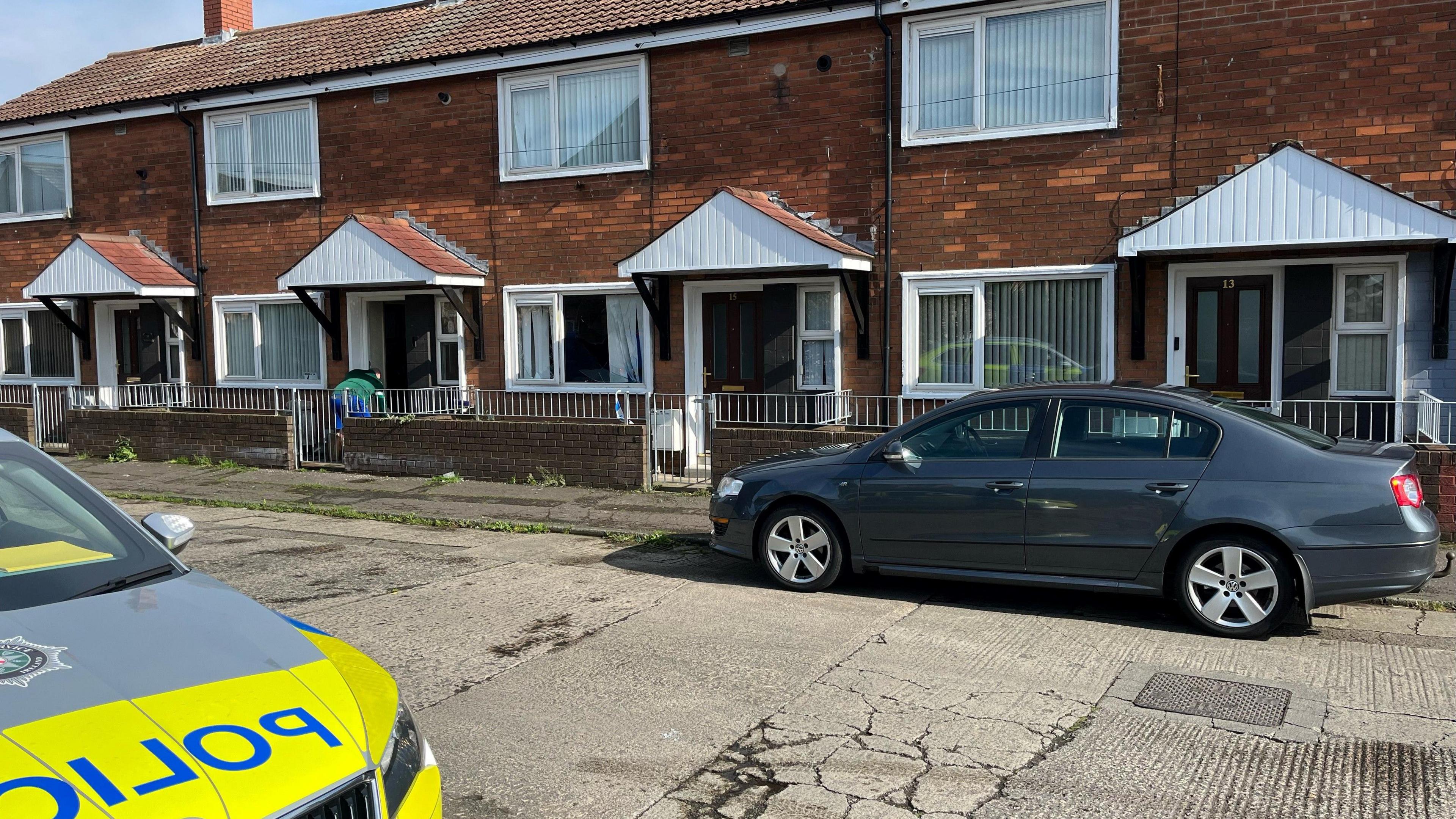 PSNI car in front of house with a person reaching down in the background and a grey car on the opposite side of the road.