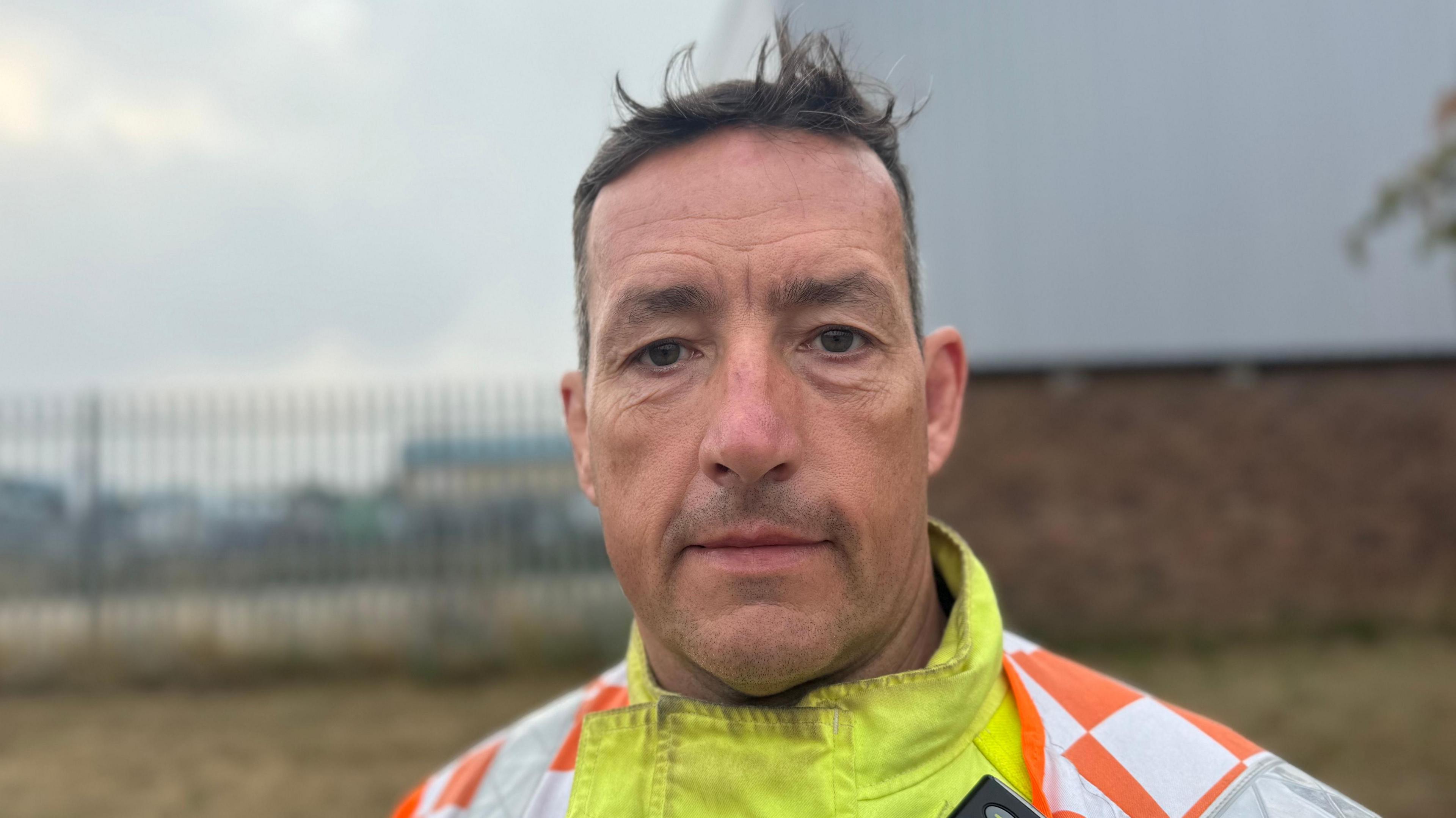 Craig McLellan with a serious expression on his face at the scene of an incident. He is wearing a high-vis jacket which is orange, white and fluorescent yellow.