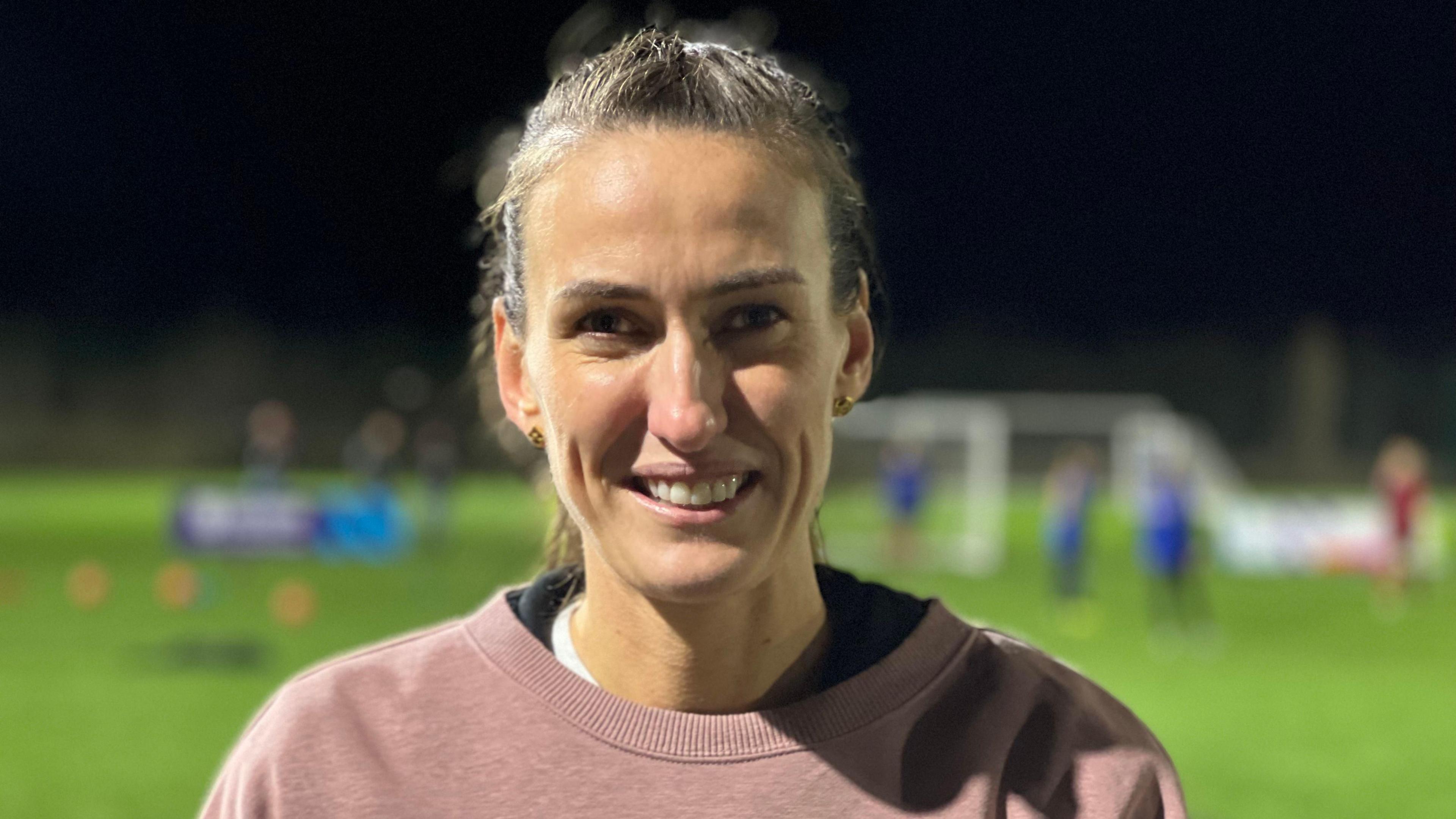 Jill Scott is standing on a football pitch as young footballers play in the background. She has long brown hair in a ponytail and is wearing a pink jumper.