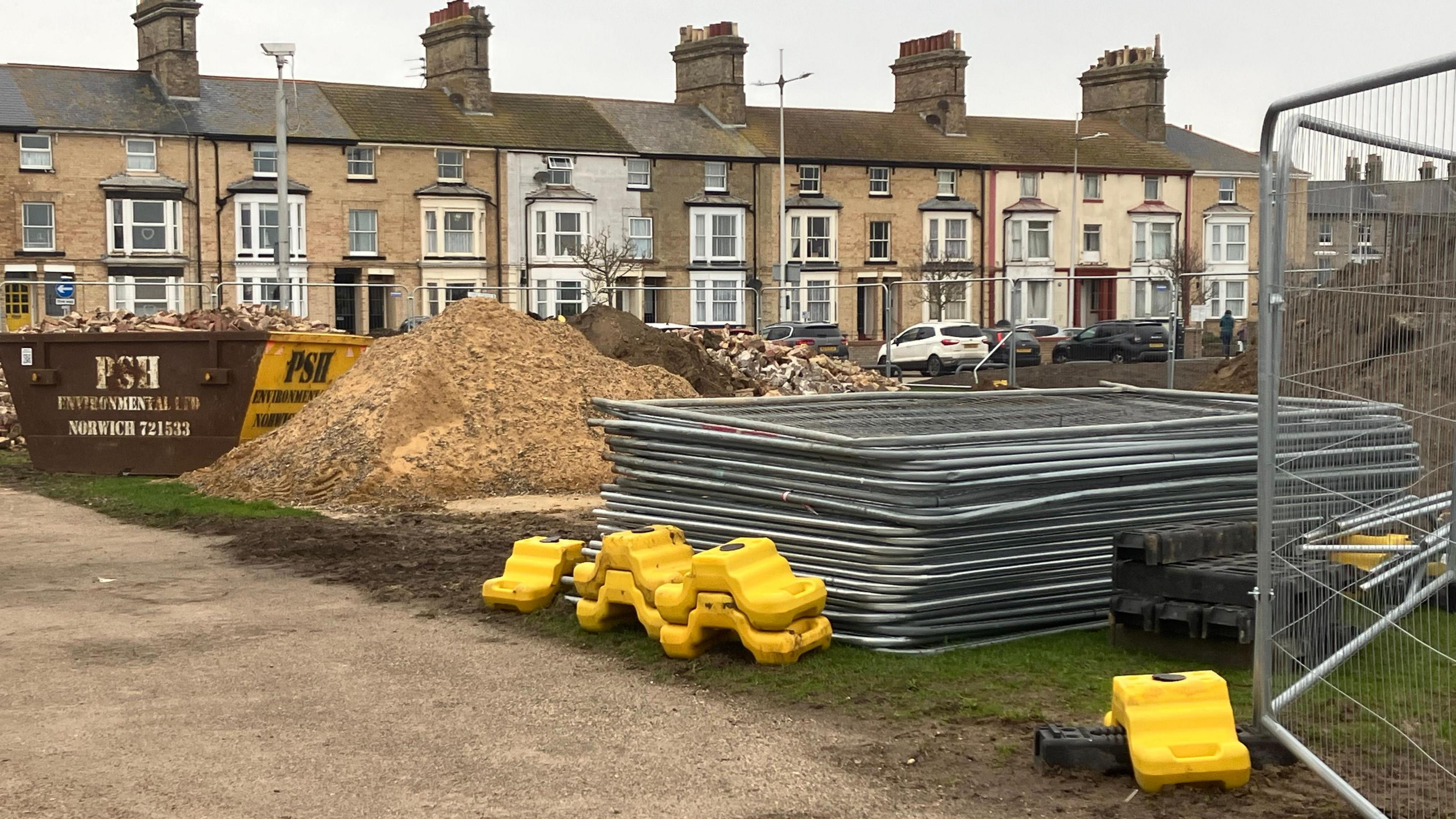 A pile of sand, a skip full of rubble and a stack of metal fencing is on a grassy area of ground in front of a row of terraced houses 