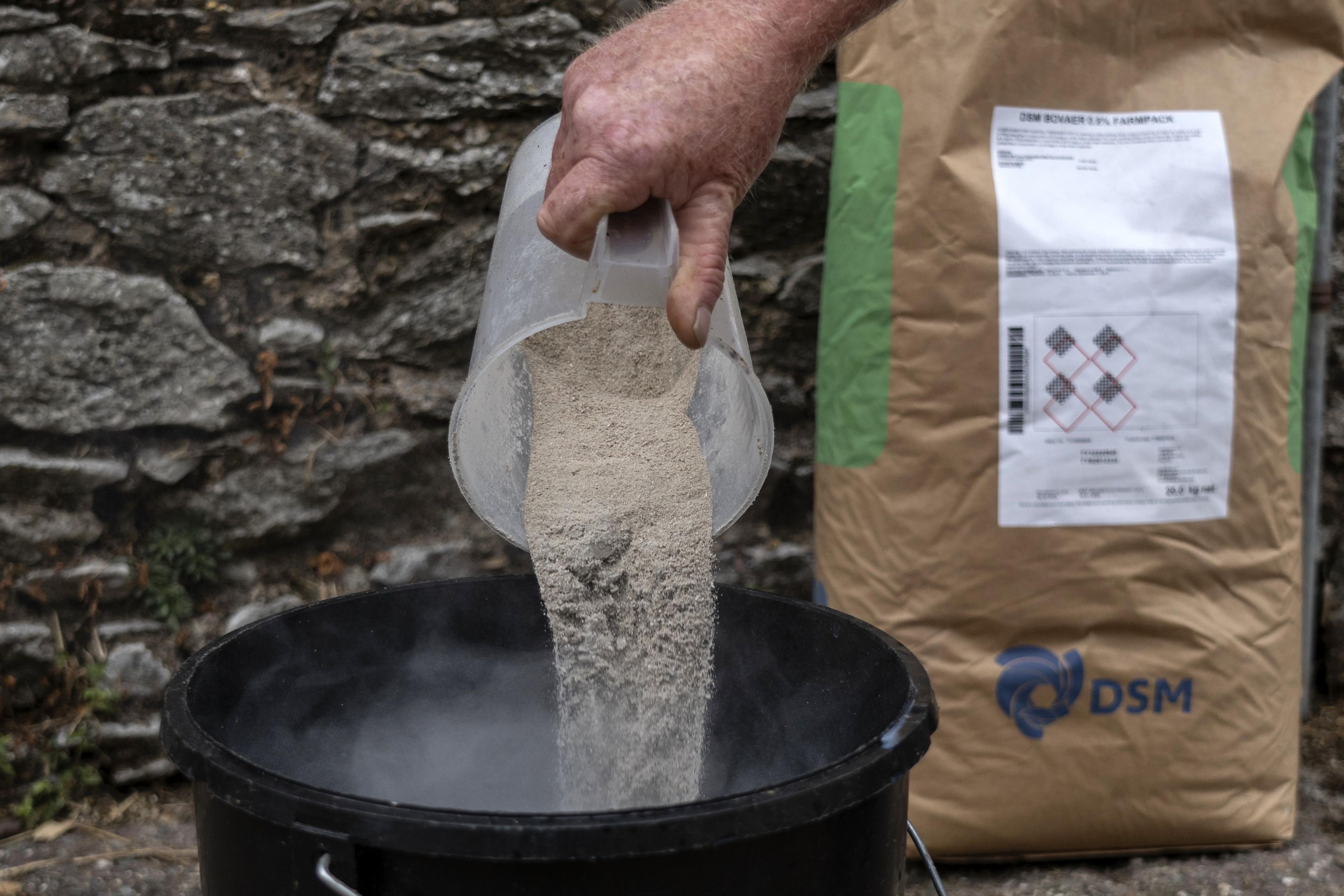 A jug of Bovaer animal feed is being poured into a bucket next to a paper bag of the product, which leans against a stone wall.