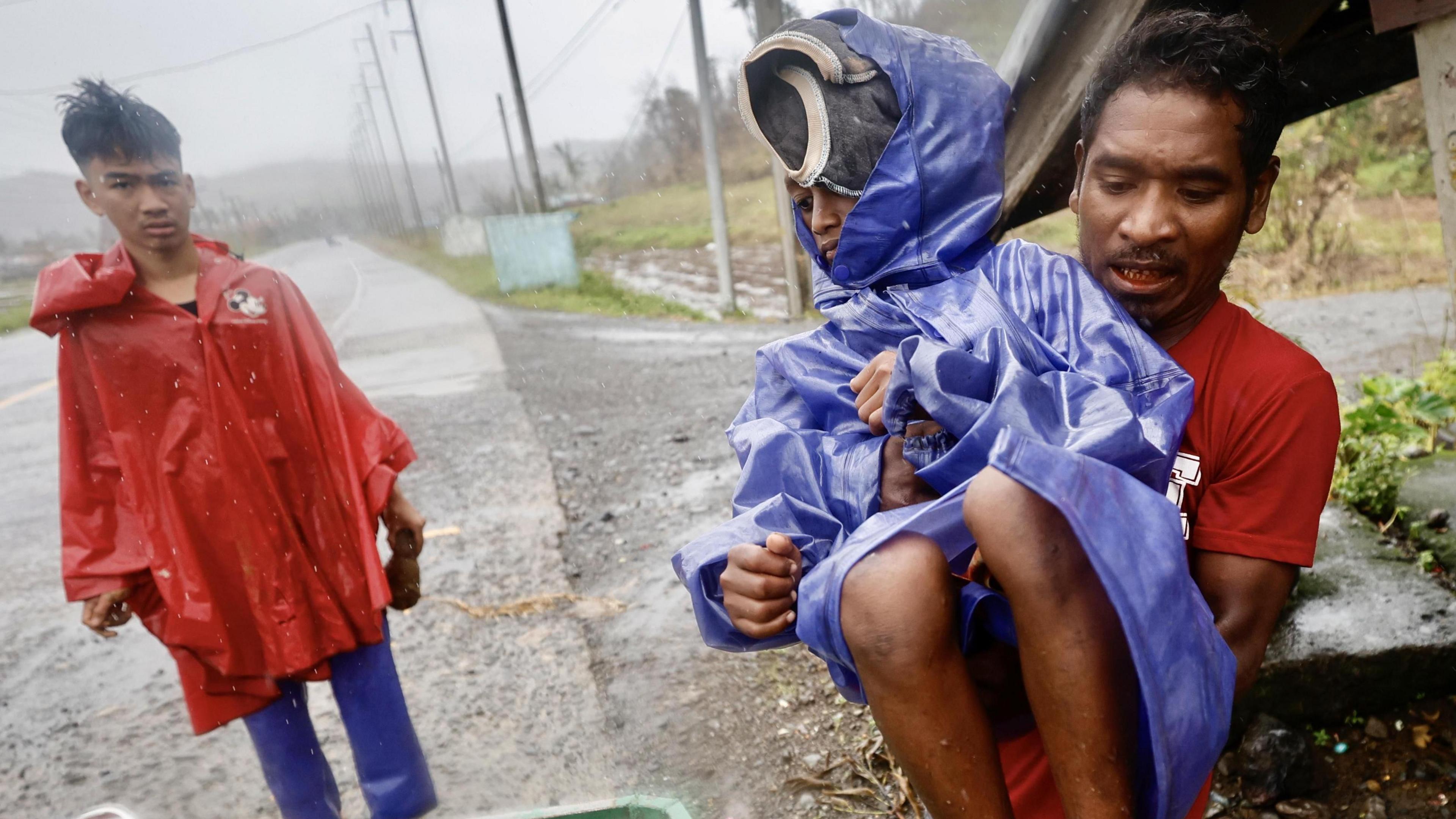 A man carries a child dressed in a purple raincoat as they prepare to evacuate from the path of a super typhoon. it's raining heavily and another youngster looks on, wearing a red rainjacket.