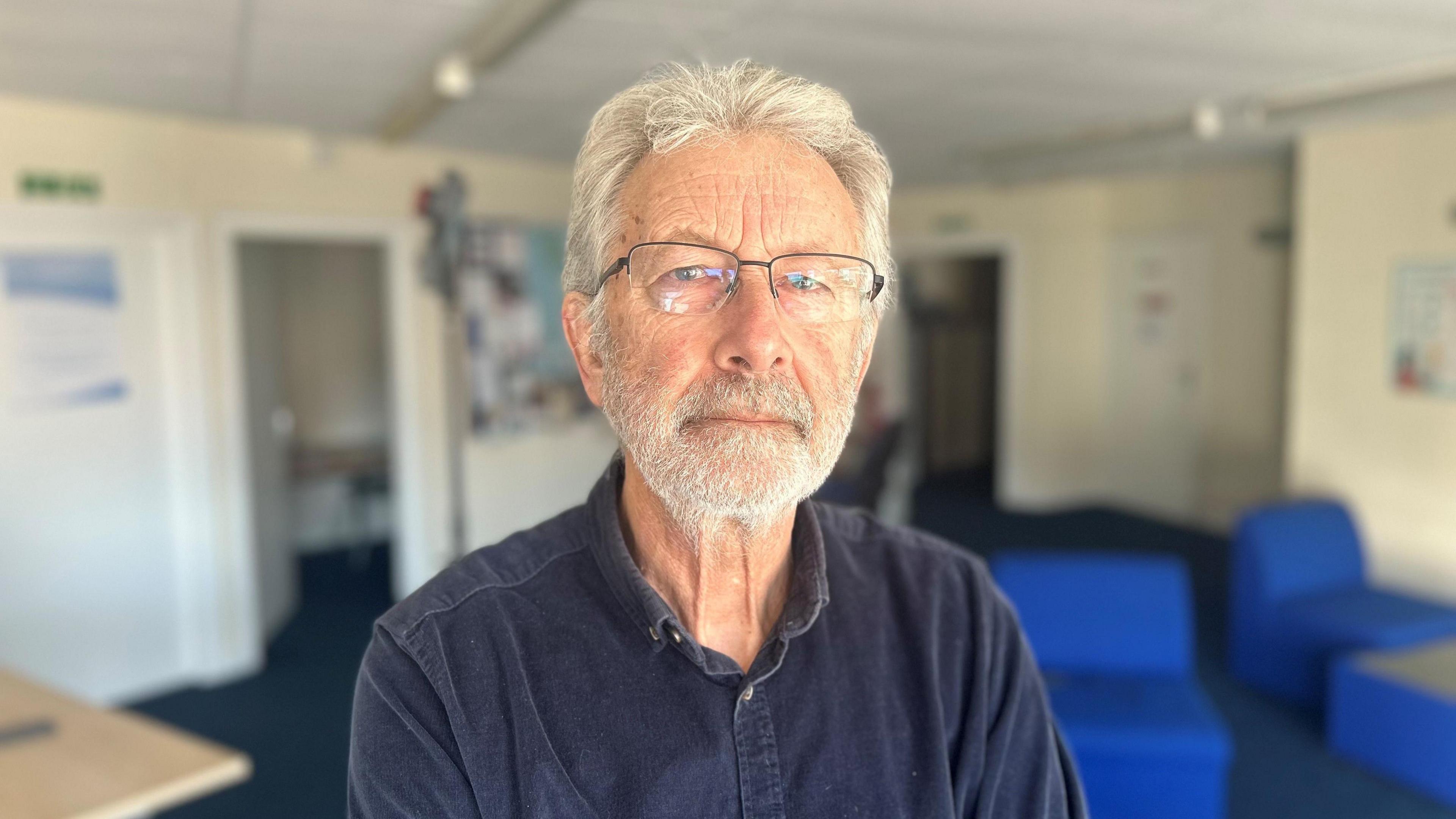 Pete Widlinski has grey hair, stubble, and a beard and moustache. He's wearing glasses and stands looking into the camera. He's in an office space with blue, out of focus, armchairs behind him. 