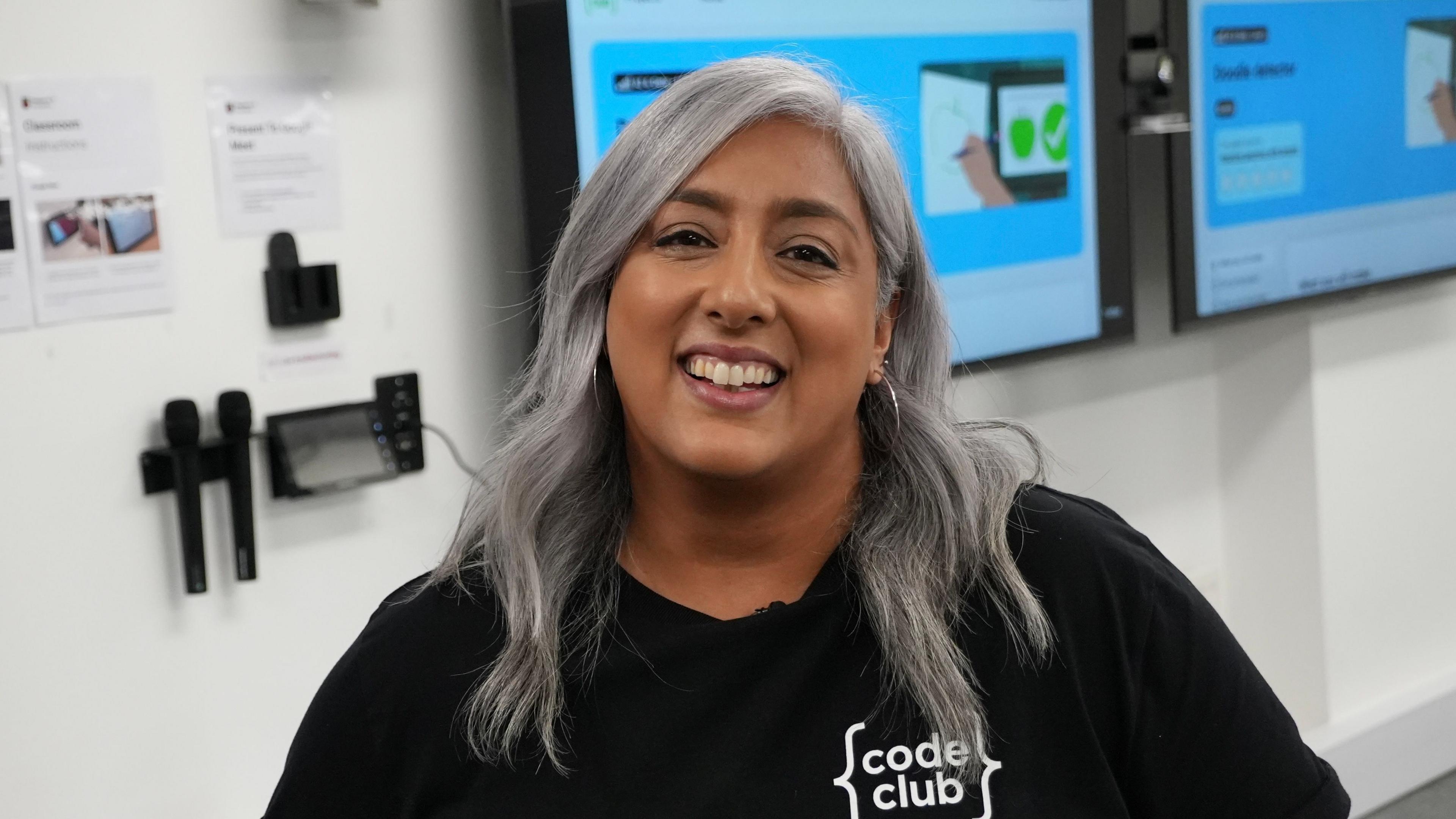 Dips Thompson is wearing a black "Code Club" t-shirt and is standing in a tech-focused learning space, likely a classroom or workshop. Behind her, two screens display coding-related content, and the walls are decorated with posters and mounted equipment. She is smiling and has long wavey grey hair and hoop earrings.