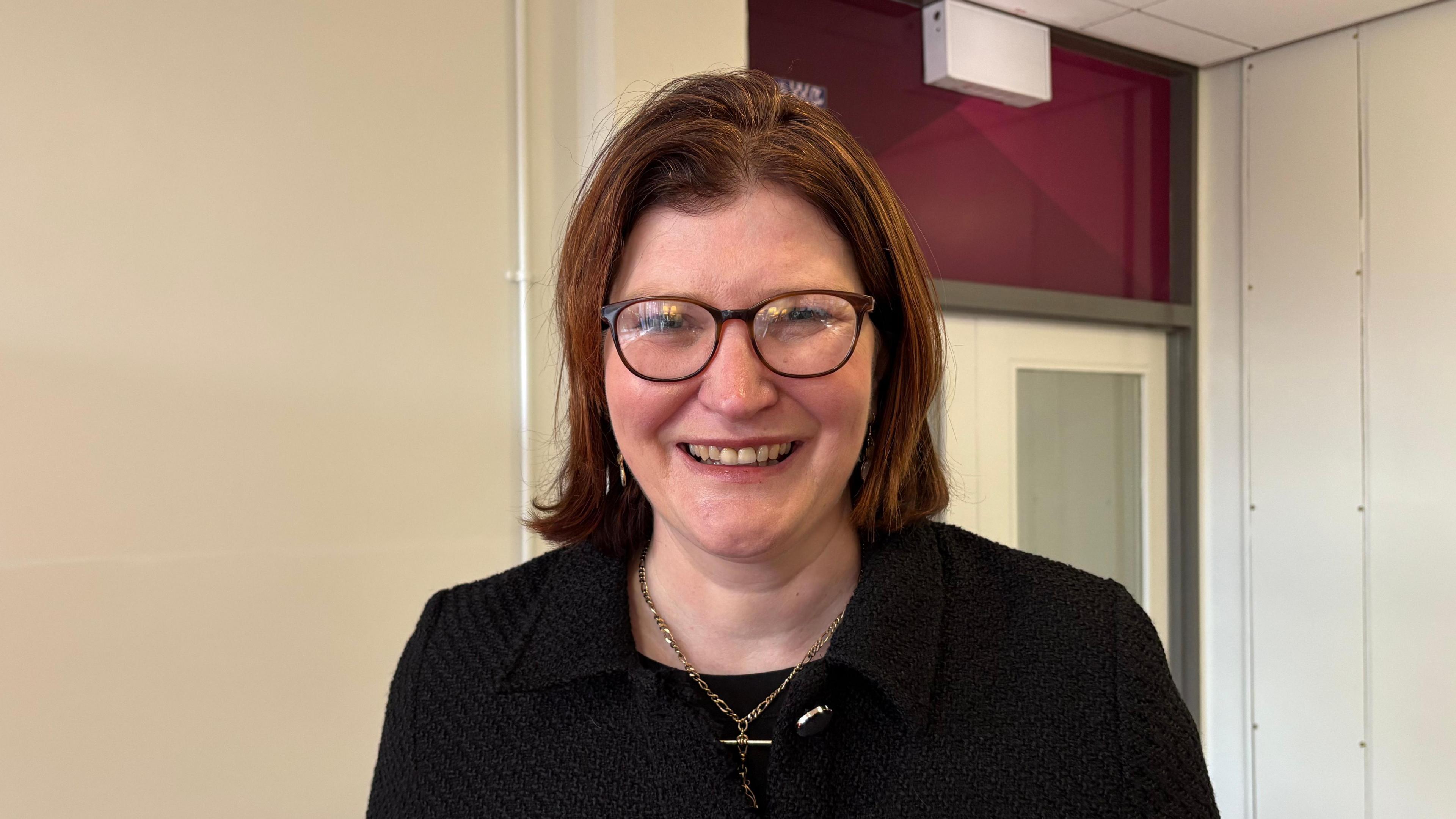 A woman with shoulder length light brown hair smiles toward the camera. She is wearing dark rimmed glasses, a black top and a black blazer. 