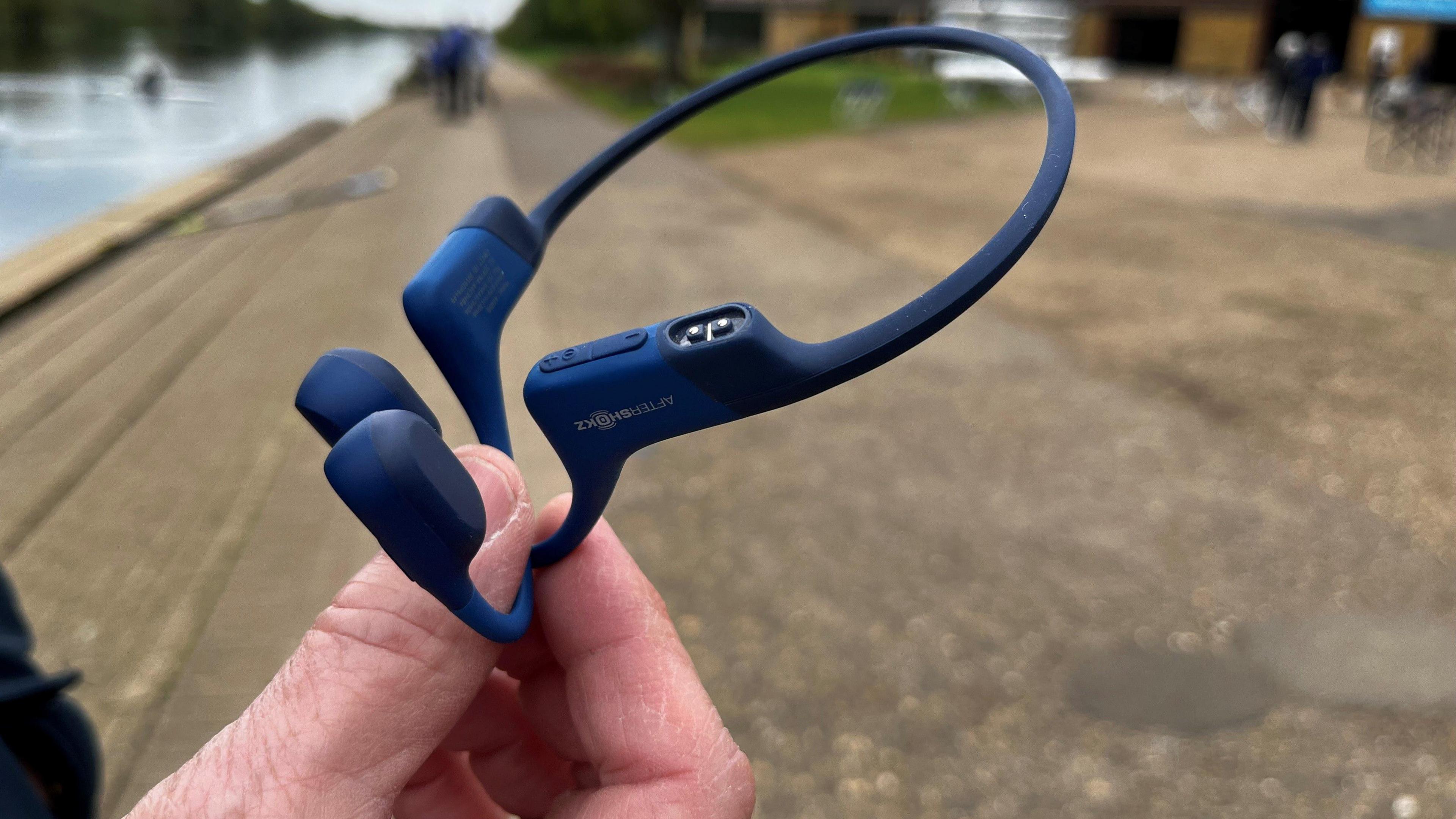 The blue and small headset is being held by someone between their fingers. In the background is the wooden decking and concrete surface of the path beside the river.