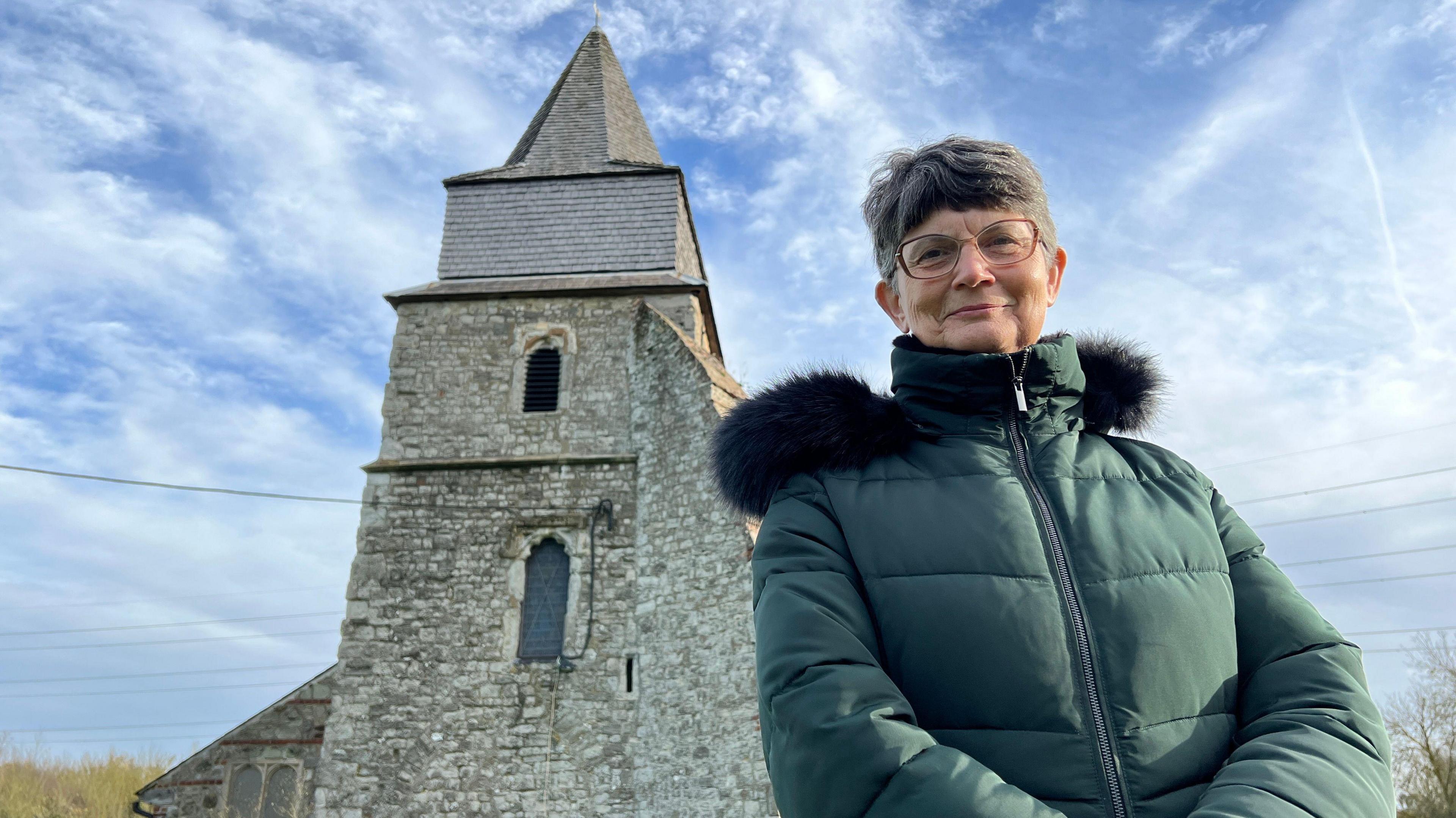 Woman with glasses wears a warm padded green jacket and stands outside a church. You can see the church tower behind her.