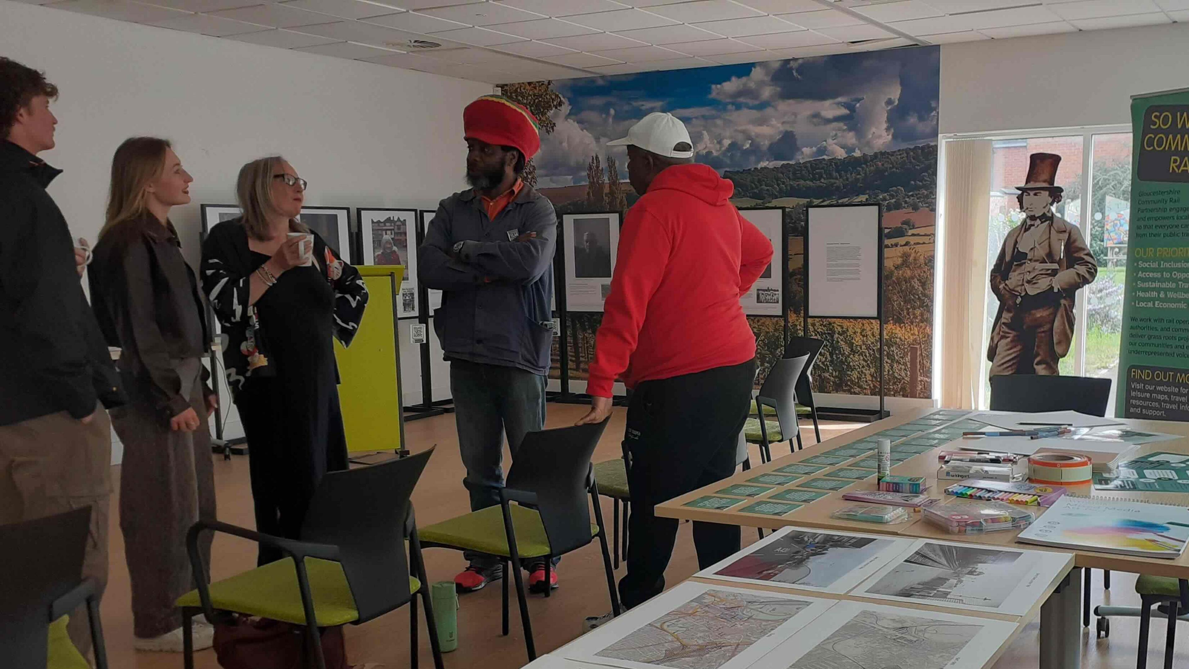 A group of people are standing next to some tables. There are photographs on the wall behind them and laid out on the tables. 