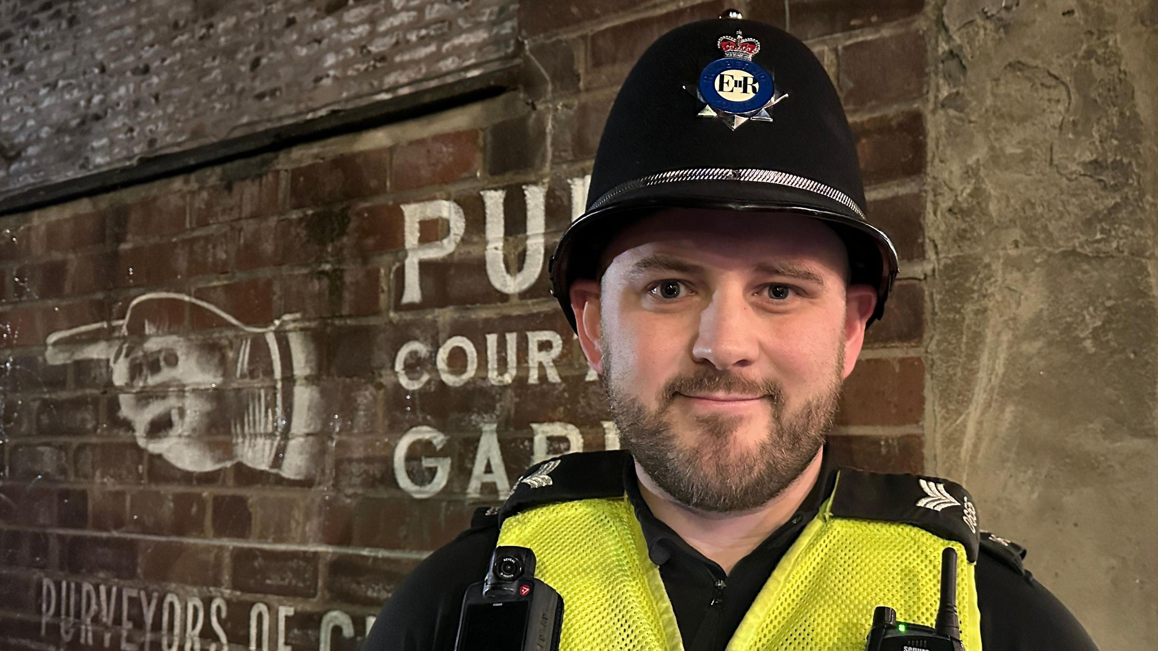 A police sergeant stands outside a pub in Beverley. He has a short brown beard and is wearing a black uniform, black helmet and a hi-vis vest. Behind him, a sign painted on a brick wall advertises a pub courtyard garden.