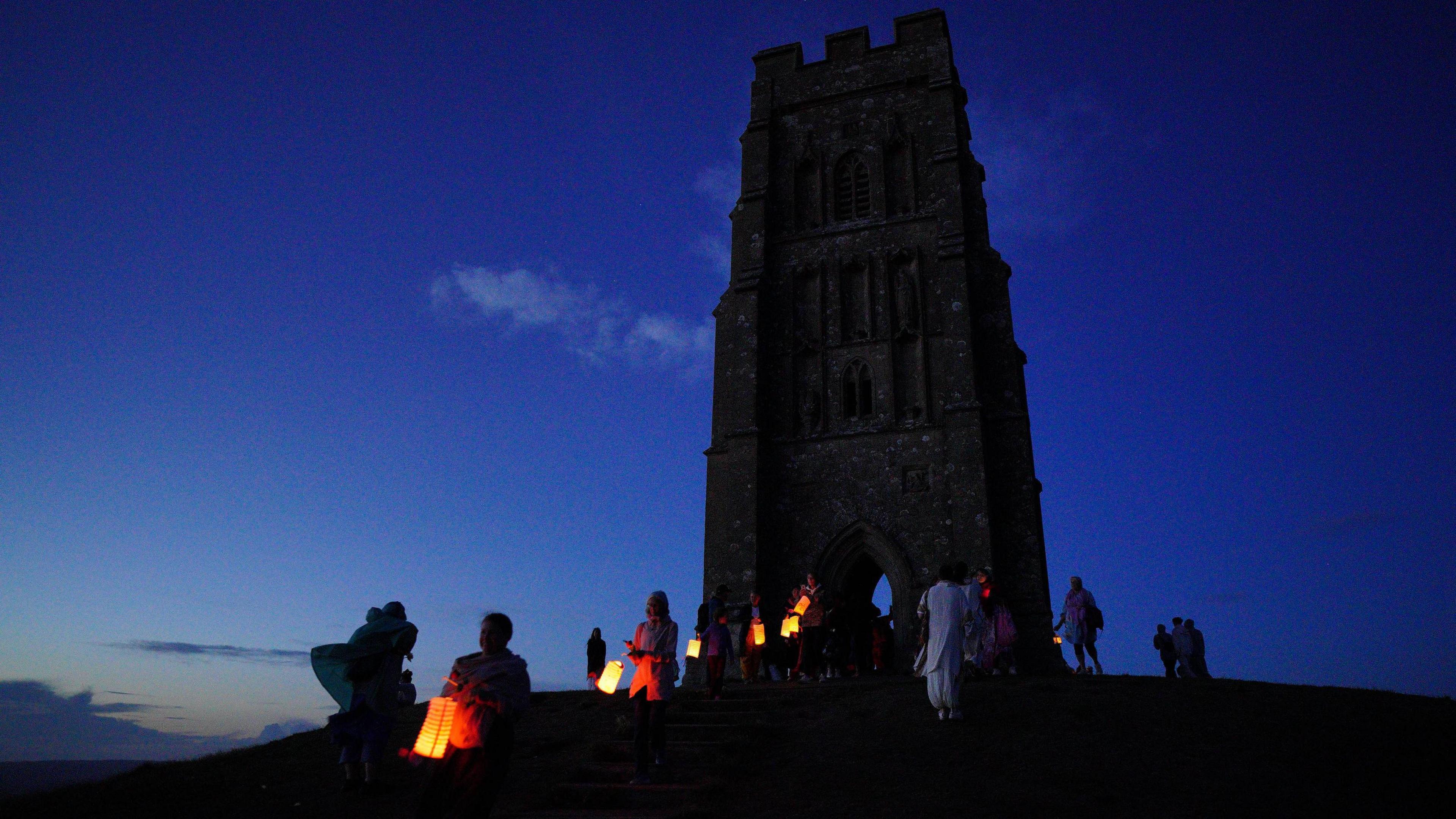 People are carrying lanterns at dawn up towards Glastonbury Tor, which is silhouetted against the dark morning sky.