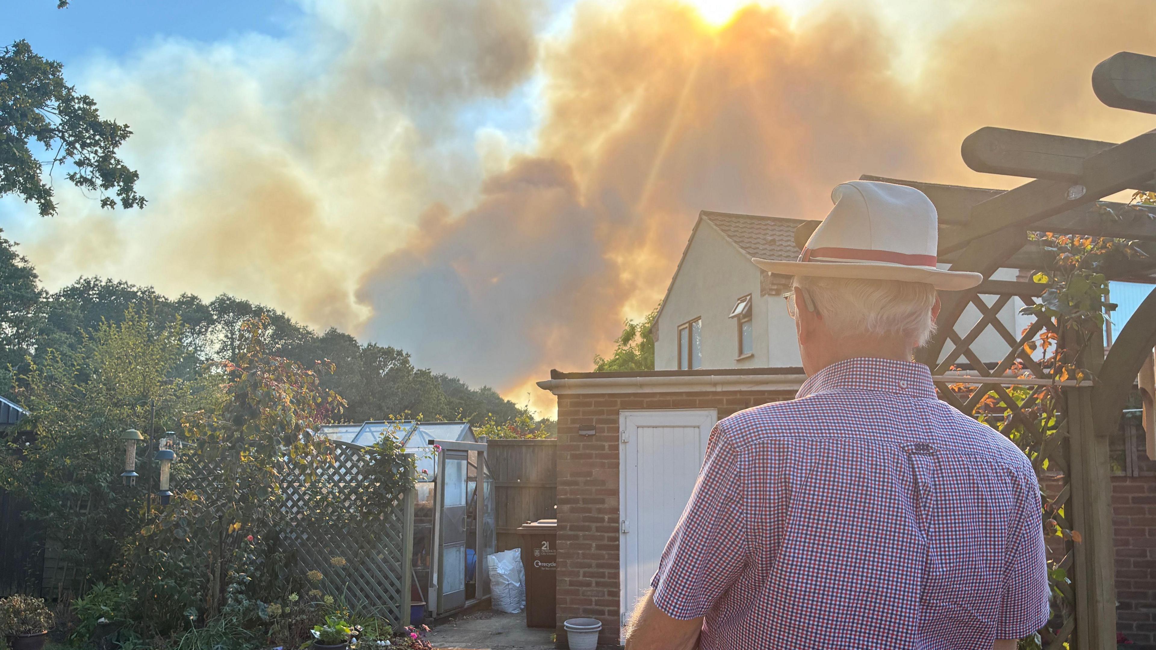 Elderly man wears a straw hat and stands with his back to the camera. He is in his garden and ahead of him are plants and a greenhouse. In the near distance, orange smoke blows in the wind.