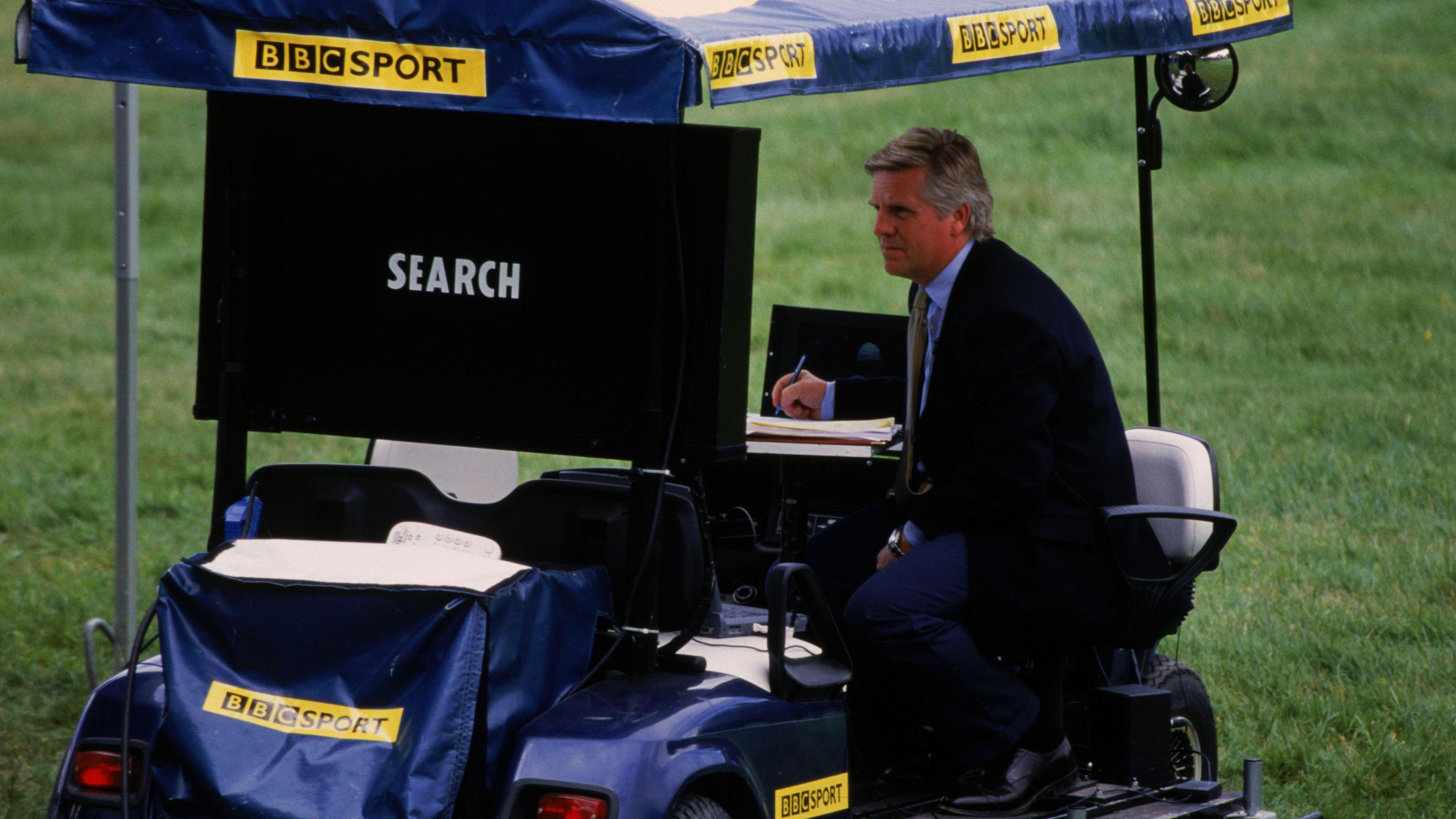 A man in a suit sitting in a golf cart which has BBC Sport branding on it.