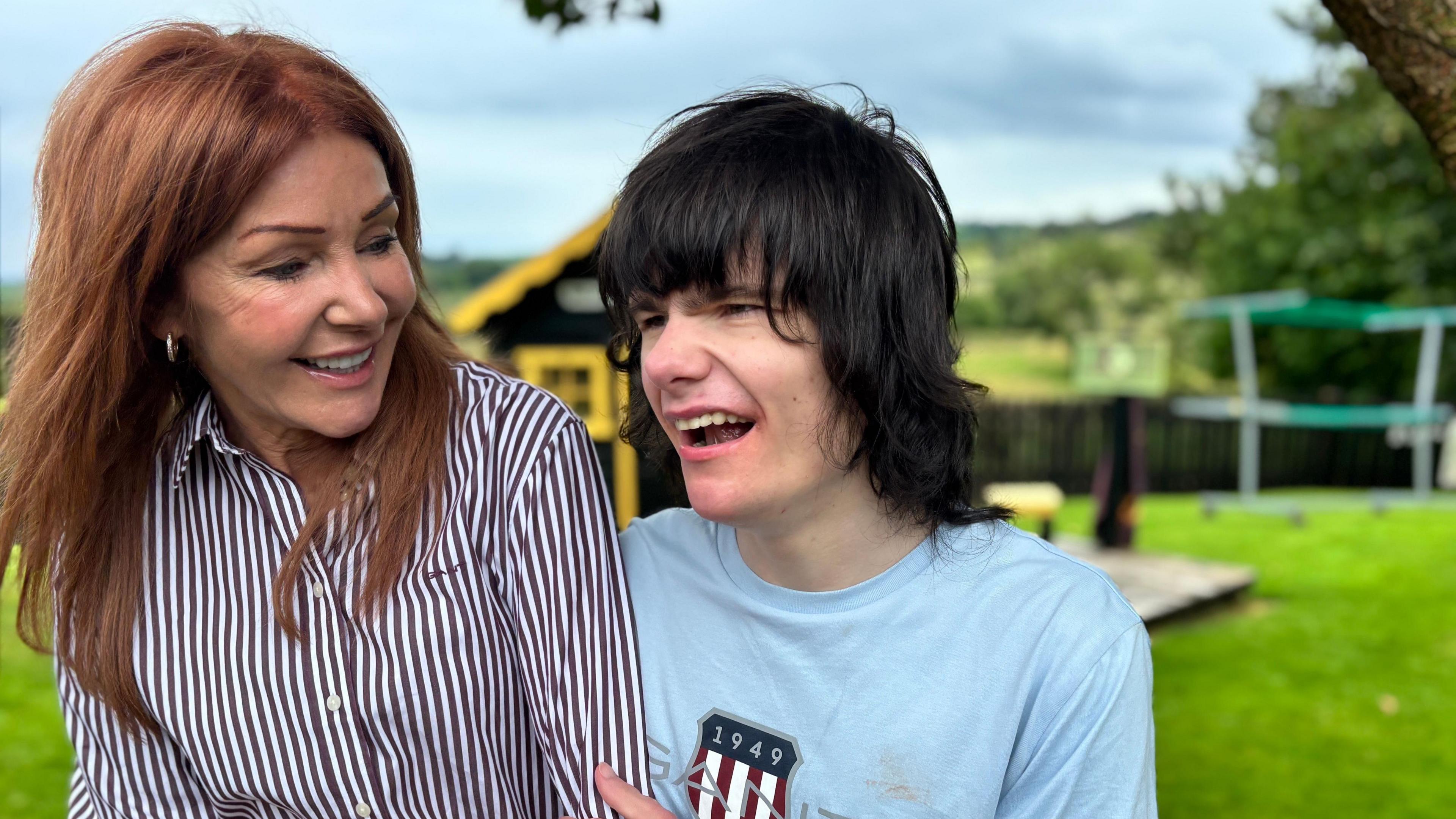 A mother and her son sit in a bright garden. The woman is smiling down fondly at her son as he holds her arm. She has long red hair that is being blown by the wind and is wearing a shirt with stripes. The young man alongside her has a big smile. 