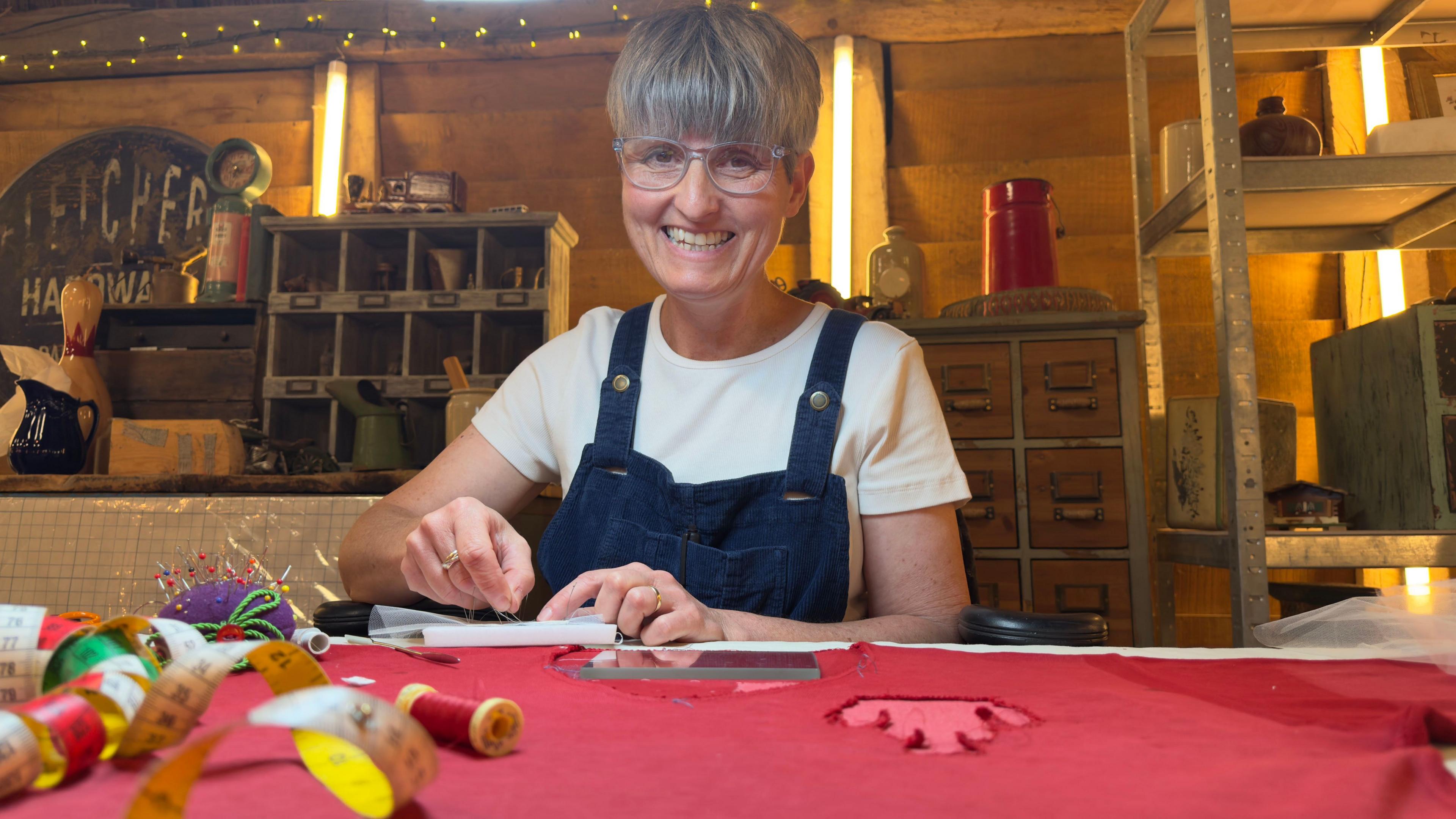 Rebecca Bissonnet, with short grey hair and wearing black dungarees over a white t-shirt, is restoring the red vintage football shirt.