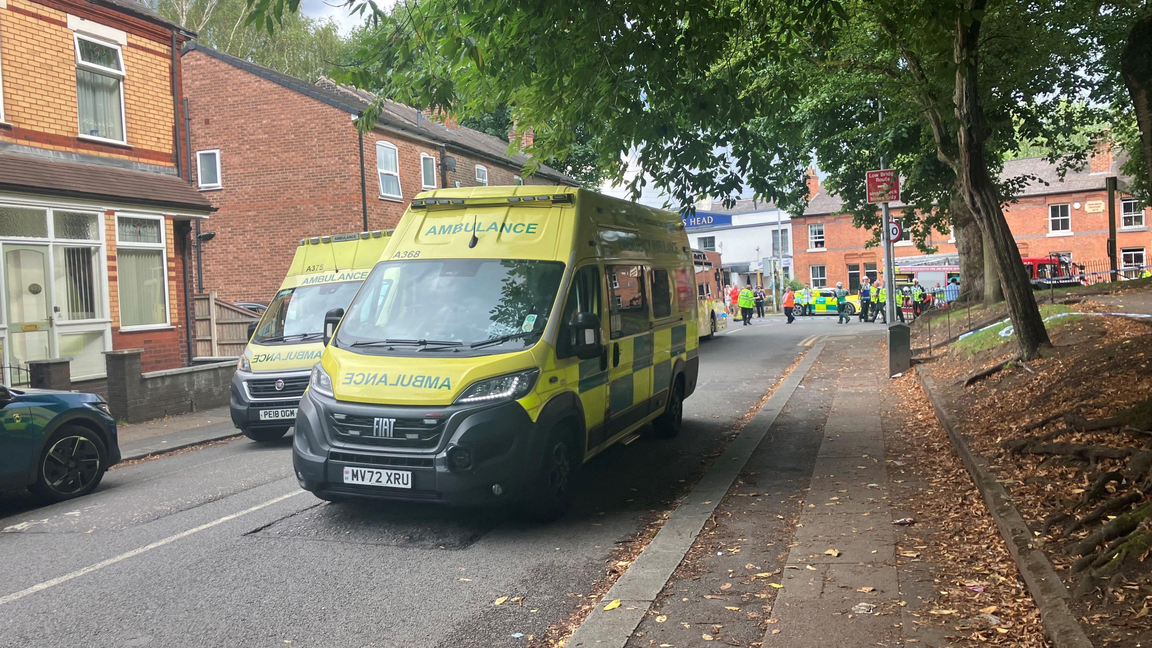 Two yellow ambulances are parked in the middle of a street. People can be seen walking along the street in the background.