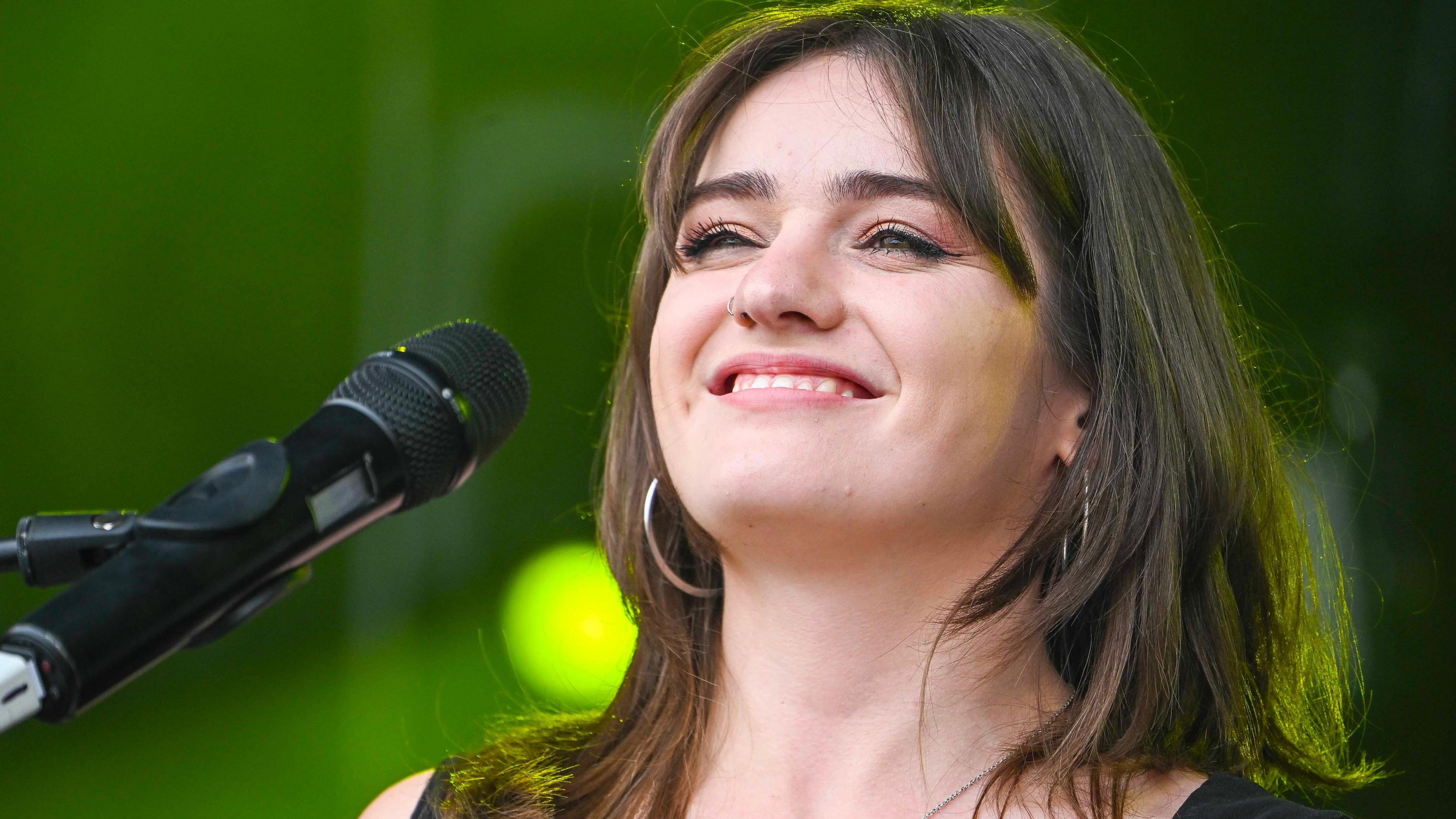 Kate Hall, a young woman with black shoulder length hair and a silver nose ring, smiles in front of a mic, with blurred green lights seen behind her.